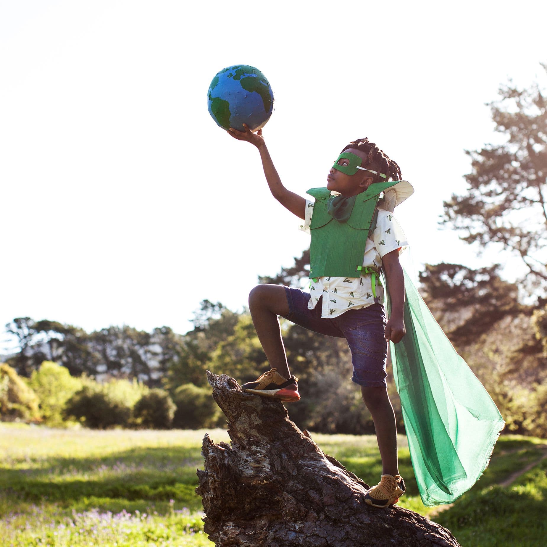 Kind mit grünem Superheldenkostüm steht auf Baumstamm und hält eine Erdkugel in die Luft in sonniger Parklandschaft