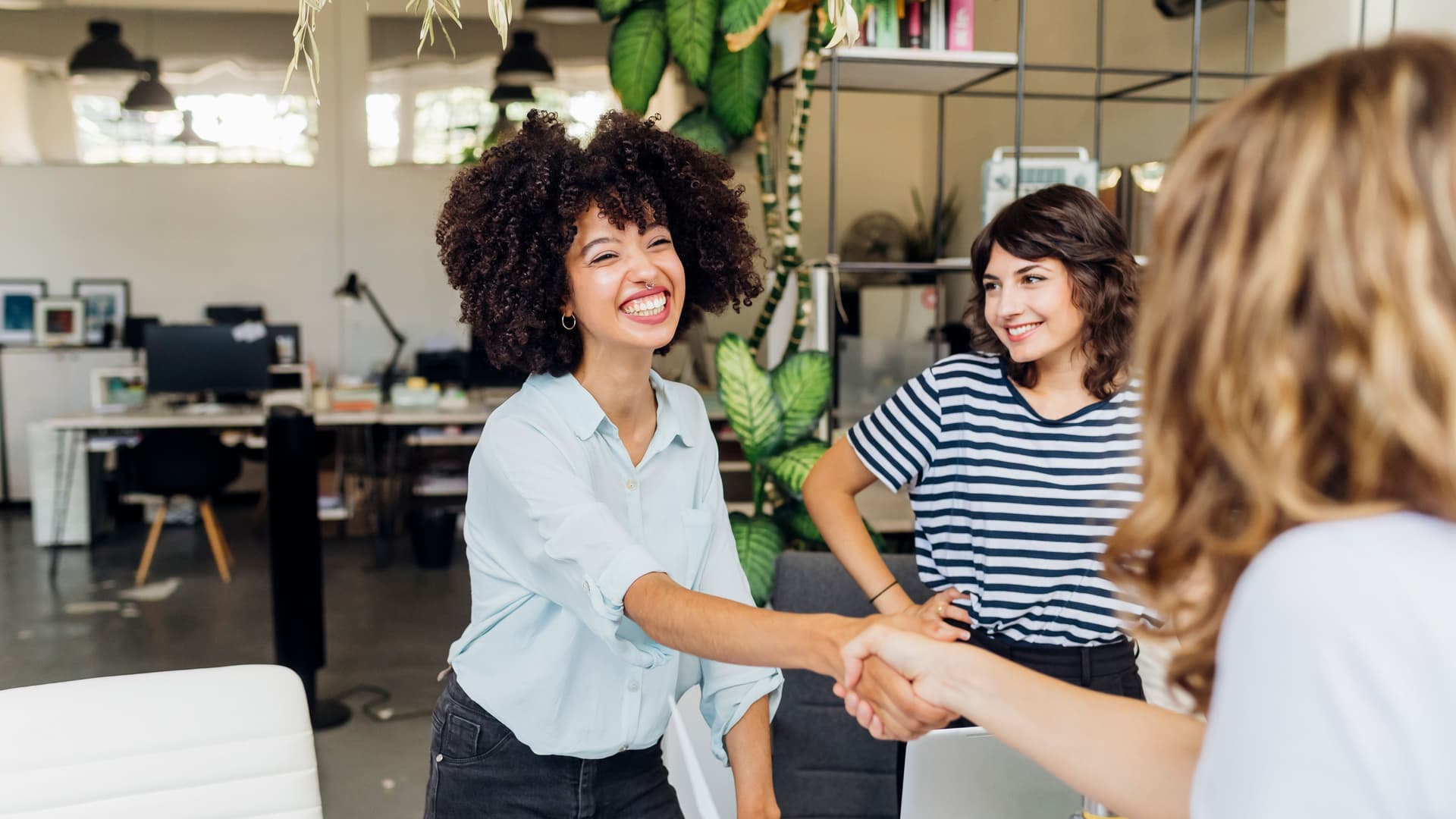 Eine junge lachende POC reicht einer anderen Person die Hand. Daneben steht eine weiter Person und lächelt.