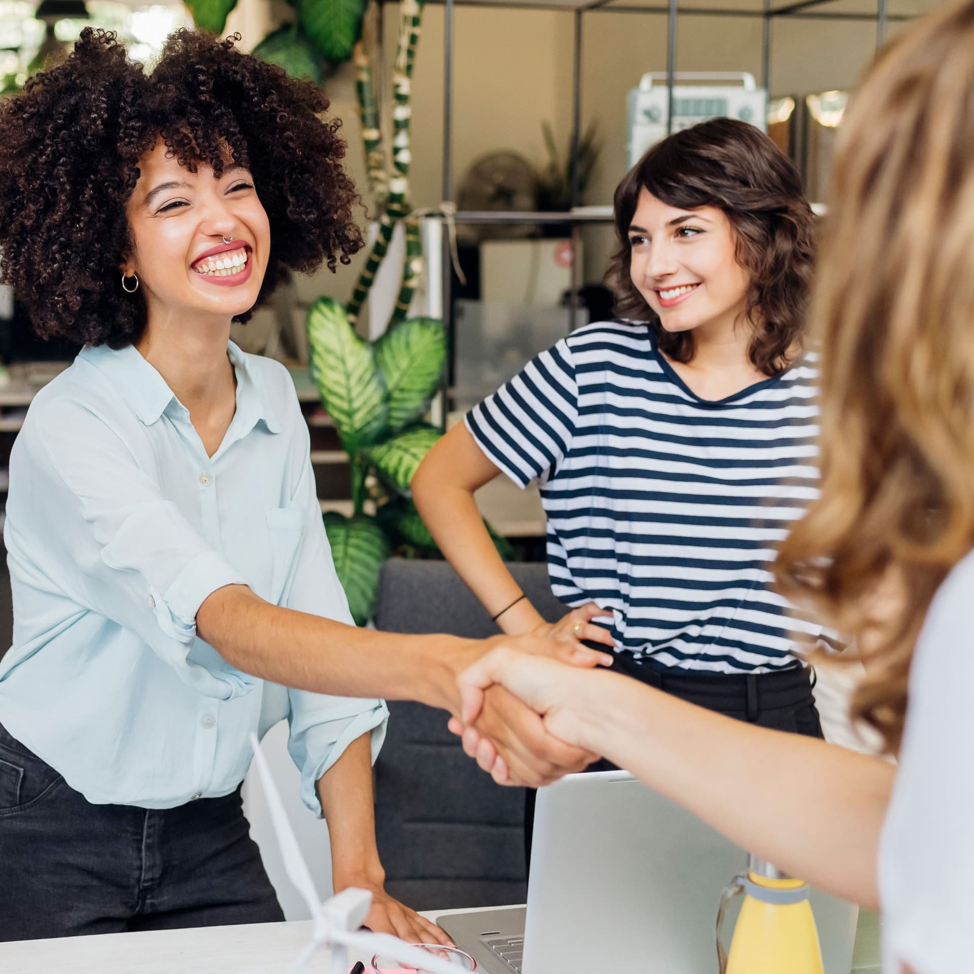 Eine junge lachende POC reicht einer anderen Person die Hand. Daneben steht eine weiter Person und lächelt.
