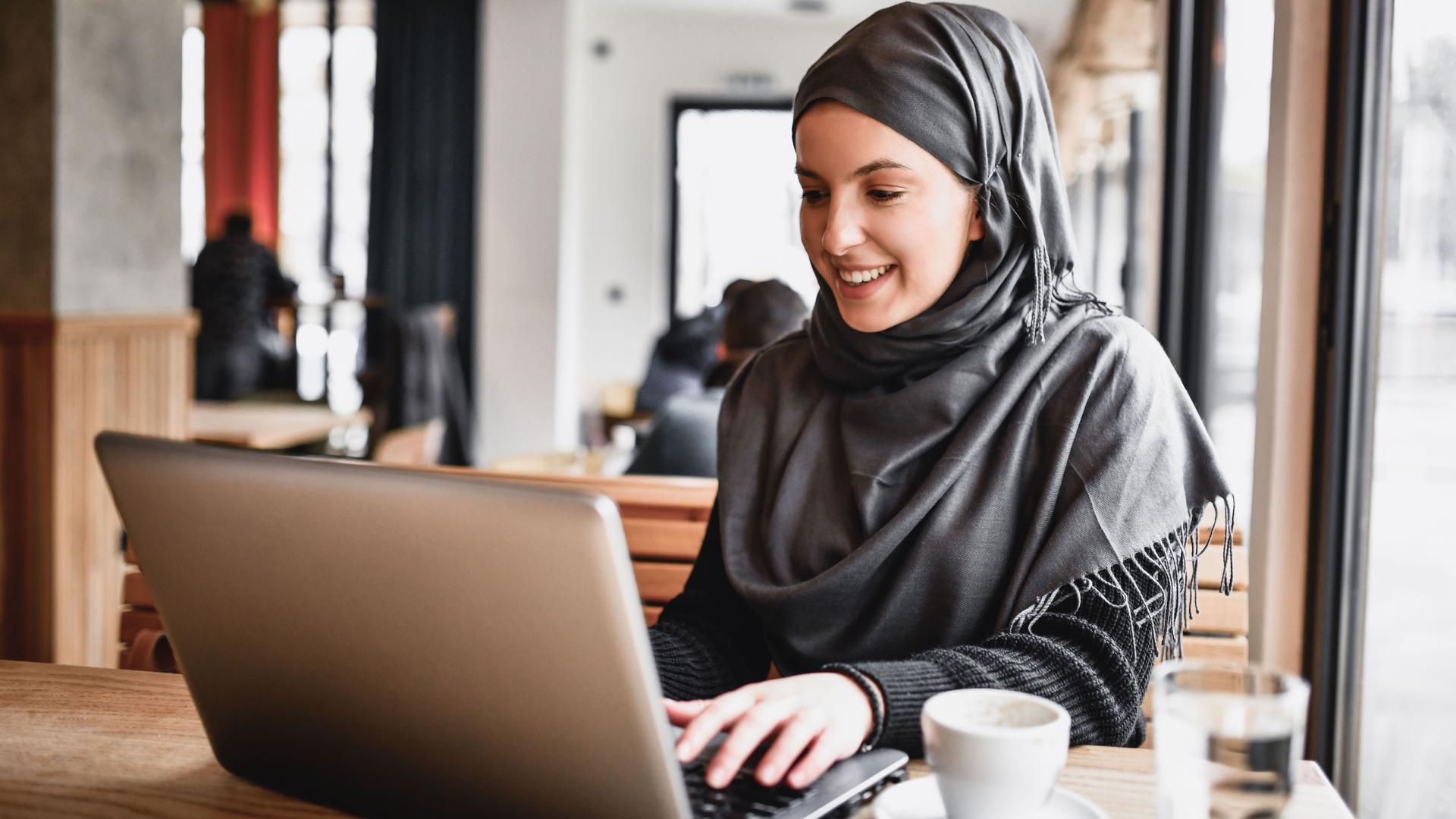 Eine junge Muslima mit grauem Kopftuch sitzt vor ihrem Laptop. Neben ihr stehen ein Kaffee und ein Glas Wasser.