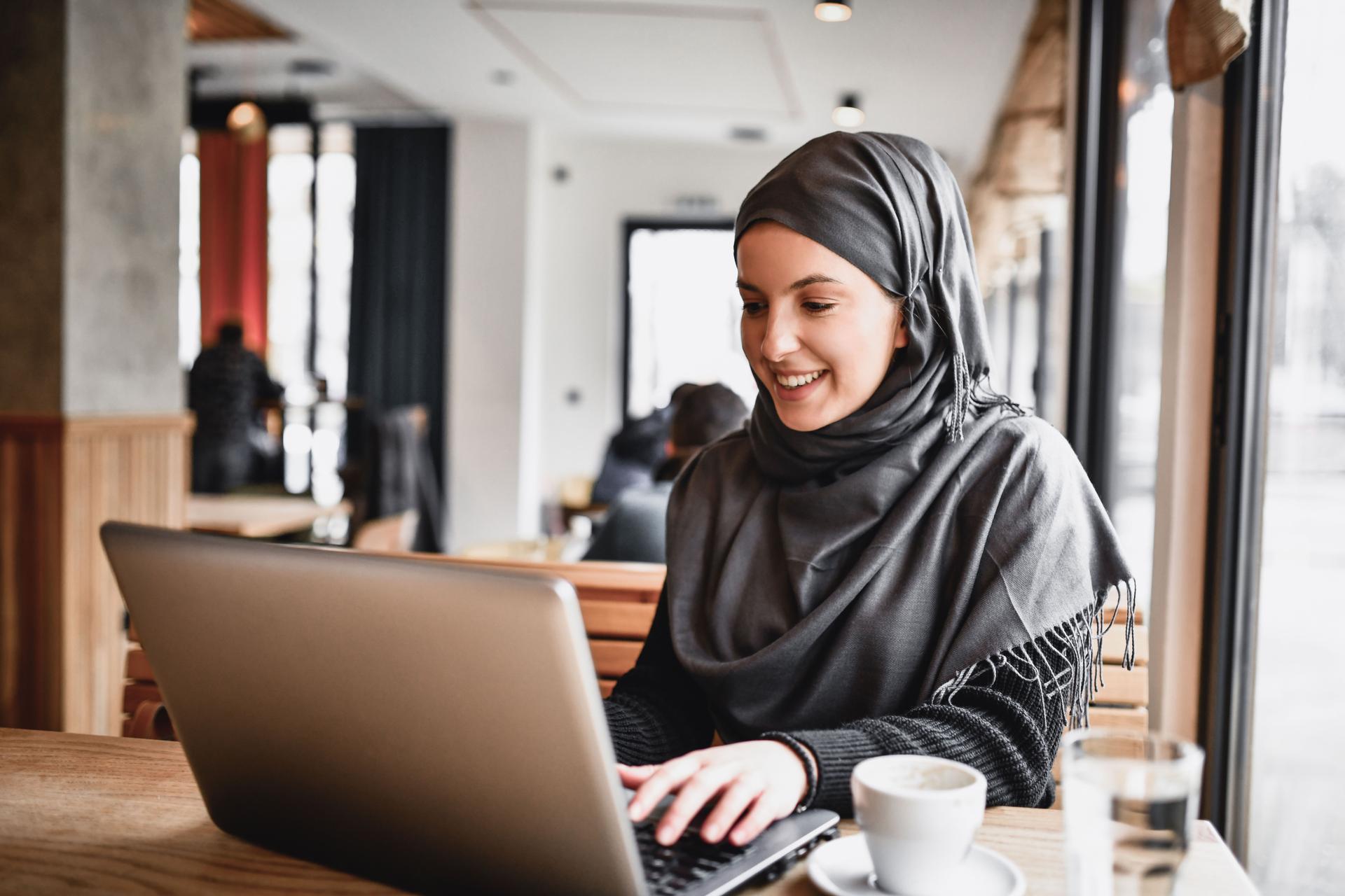 Eine junge Muslima mit grauem Kopftuch sitzt vor ihrem Laptop. Neben ihr stehen ein Kaffee und ein Glas Wasser.