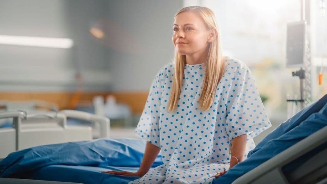 Eine Frau sitzt im Krankenhaus auf einem Bett in einem Krankenhaushemd. Sie lächelt dabei. Im Hintergrund sind weitere Betten und ein Glasfenster zu erkennen.
