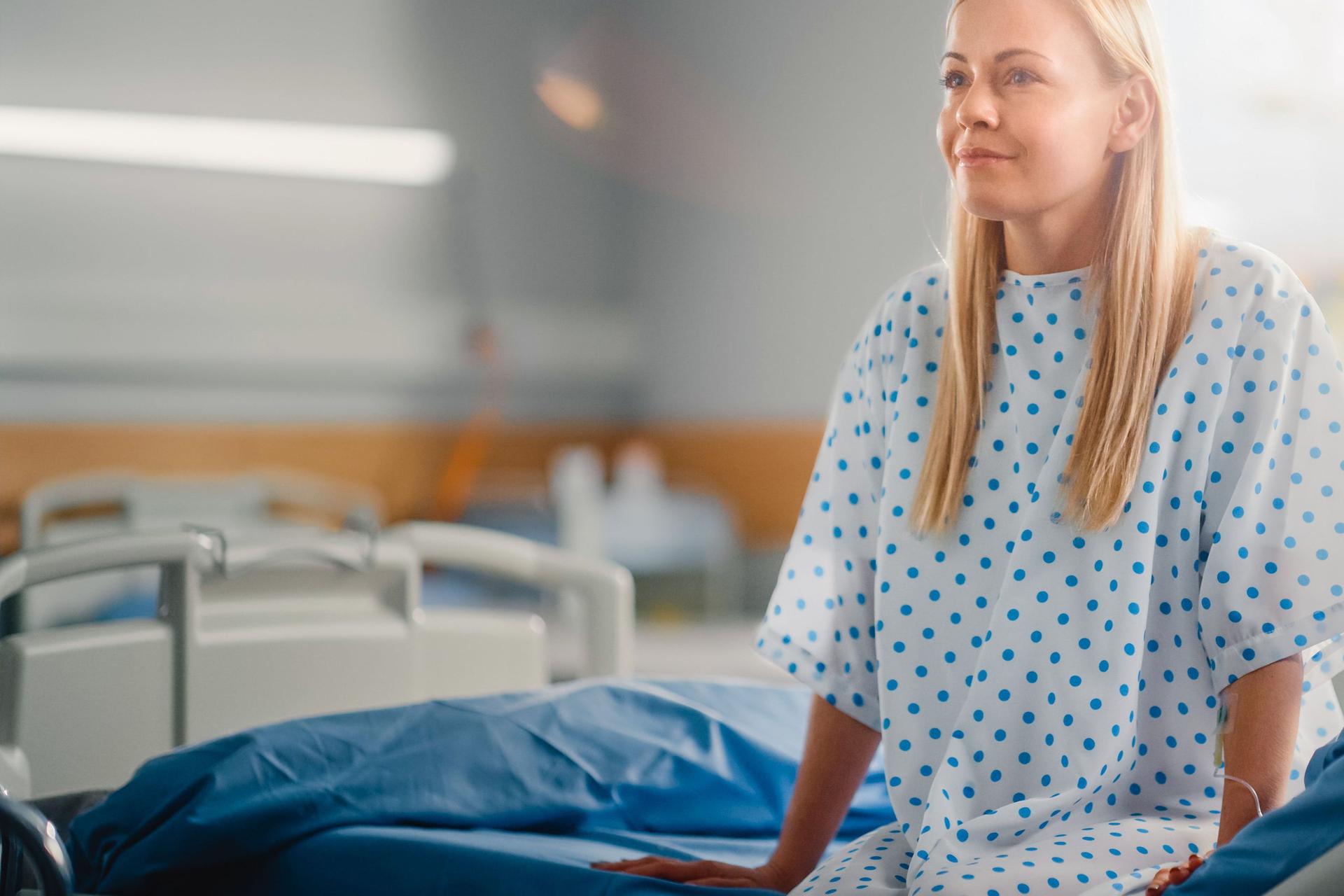 Eine Frau sitzt im Krankenhaus auf einem Bett in einem Krankenhaushemd. Sie lächelt dabei. Im Hintergrund sind weitere Betten und ein Glasfenster zu erkennen.