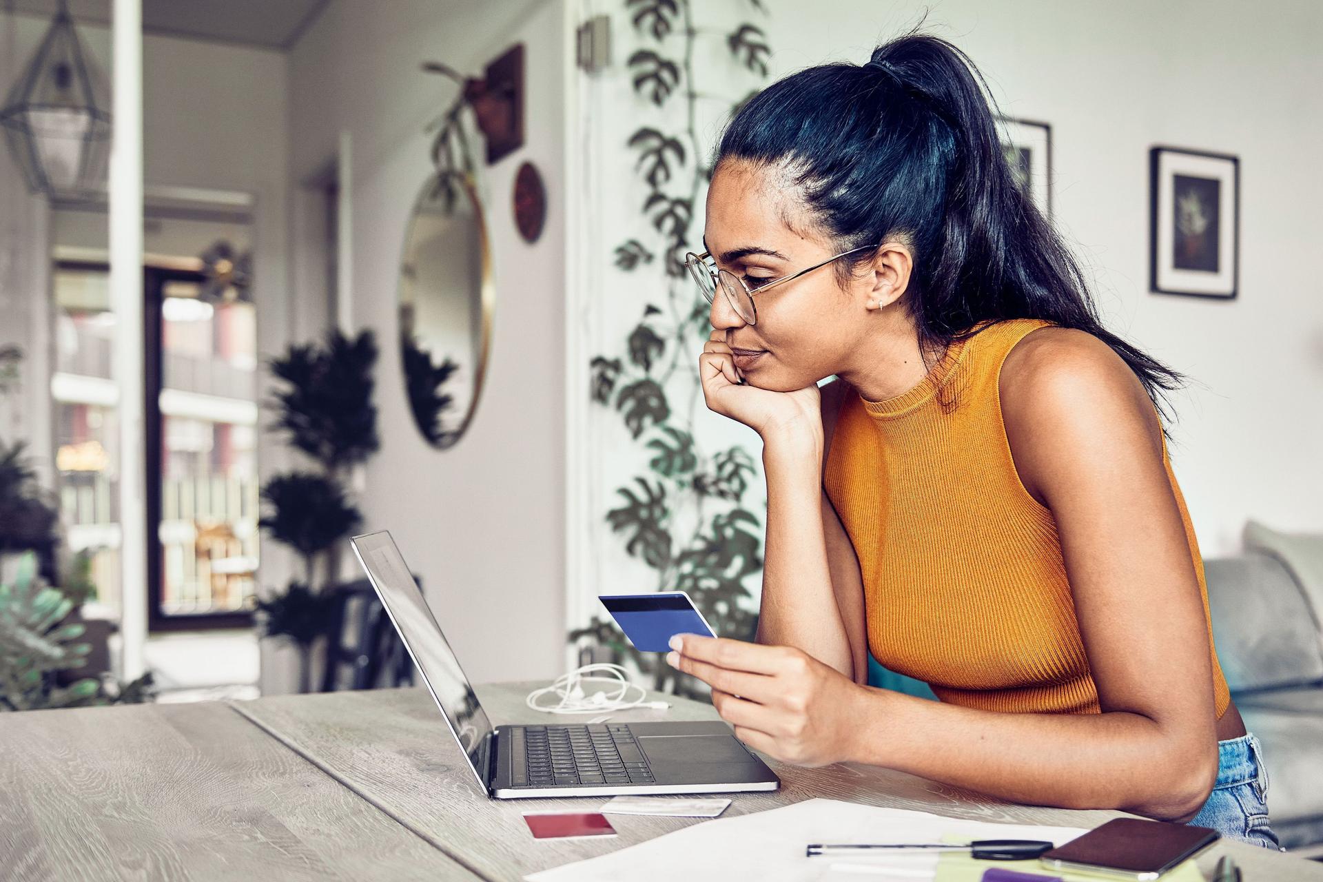 Junge Frau sitzt im Wohnzimmer vor einem Laptop mit einer Bankkarte in der Hand.