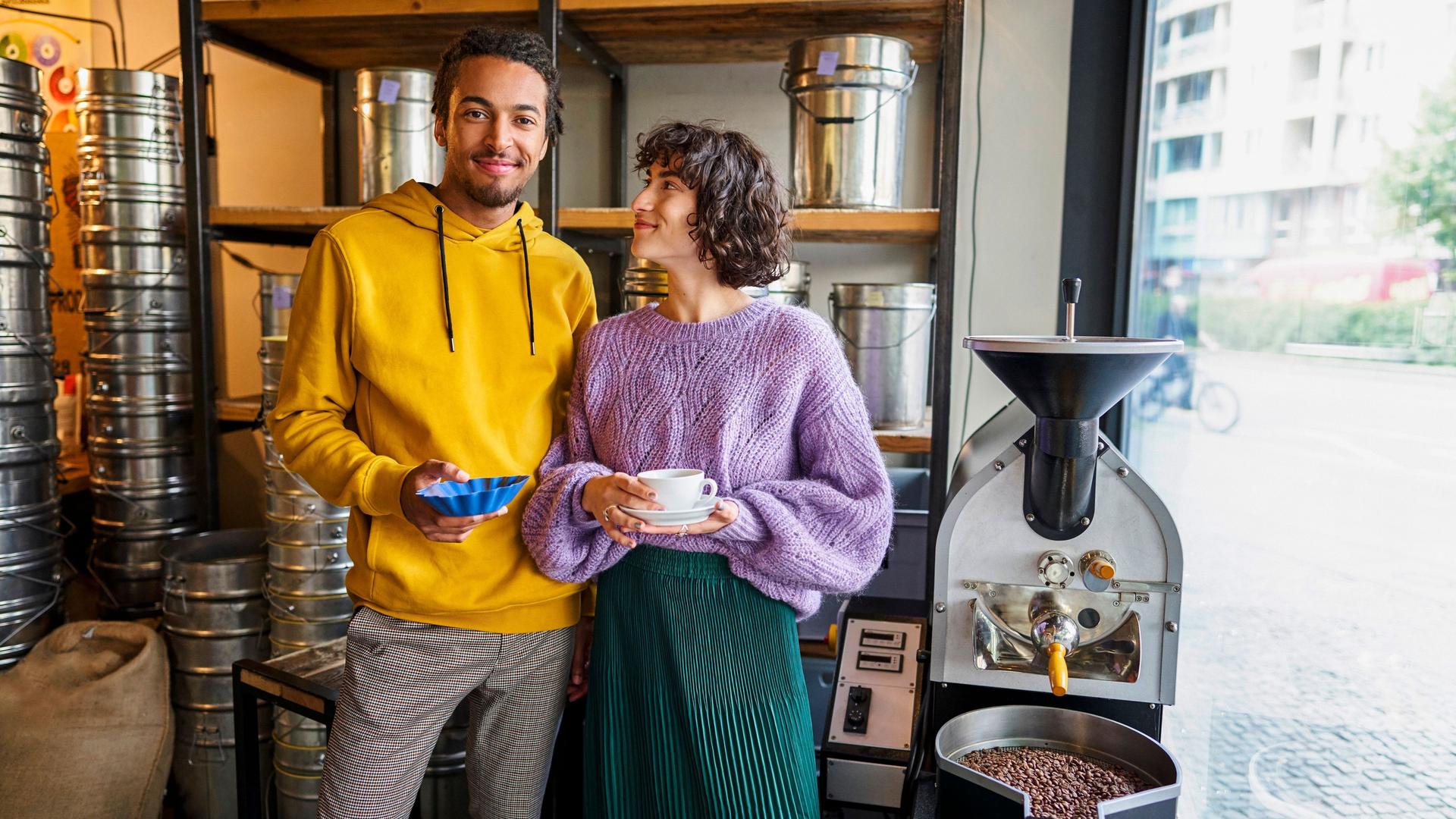 Eine junge Frau und ein junger Mann stehen in einer Kaffeerösterei in einem Ladengeschäft. Sie hat eine Tasse in der Hand und er eine Schale mit Kaffeebohnen.