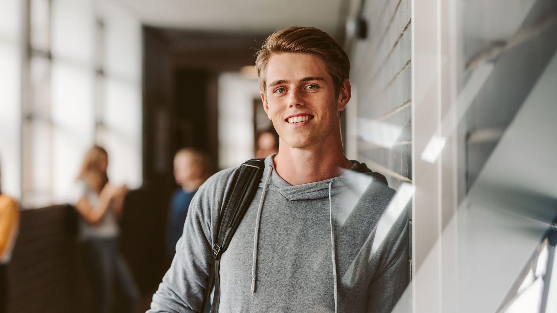 Ein sportlich gekleideter  junger Mann lehnt an einer Wand und lächelt in die Kamera. Im Hintergund stehen weitere Schüler in einem Flur.