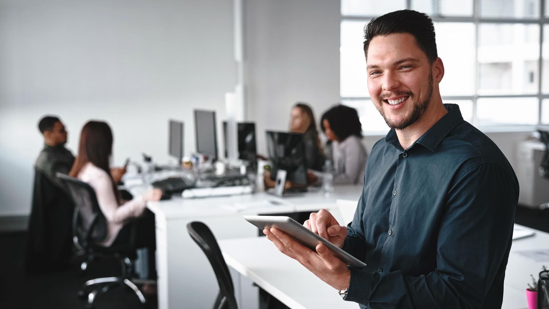 Lachender junger Geschäftsmann mit digitalem Tablet in der Hand, der in die Kamera blickt. Im Hintergrund arbeiteten seine Kollegen in einem Büro am Computer.