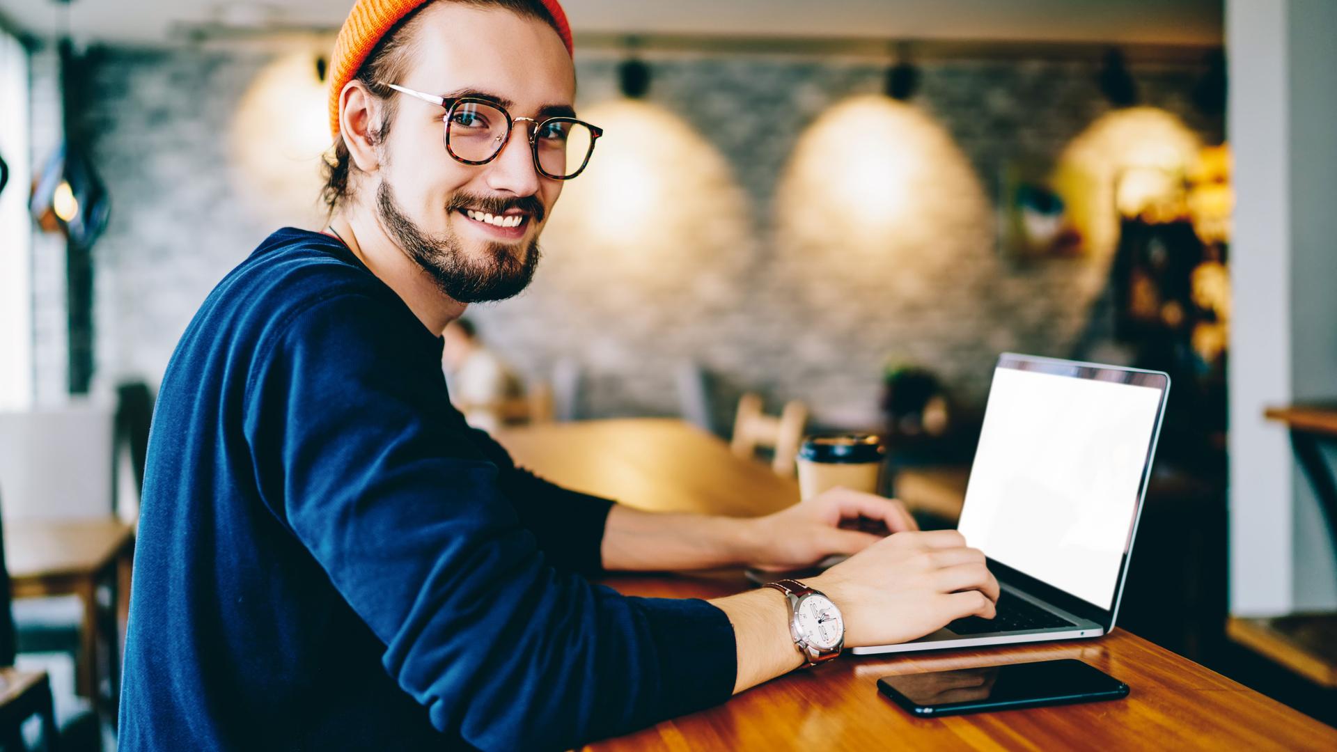Junger Mann mit Brille und Mütze guckt lächelnd in die Kamera. Er ist in einem Cafe und auf dem Tisch vor ihm ist ein Laptop und ein Smartphone.