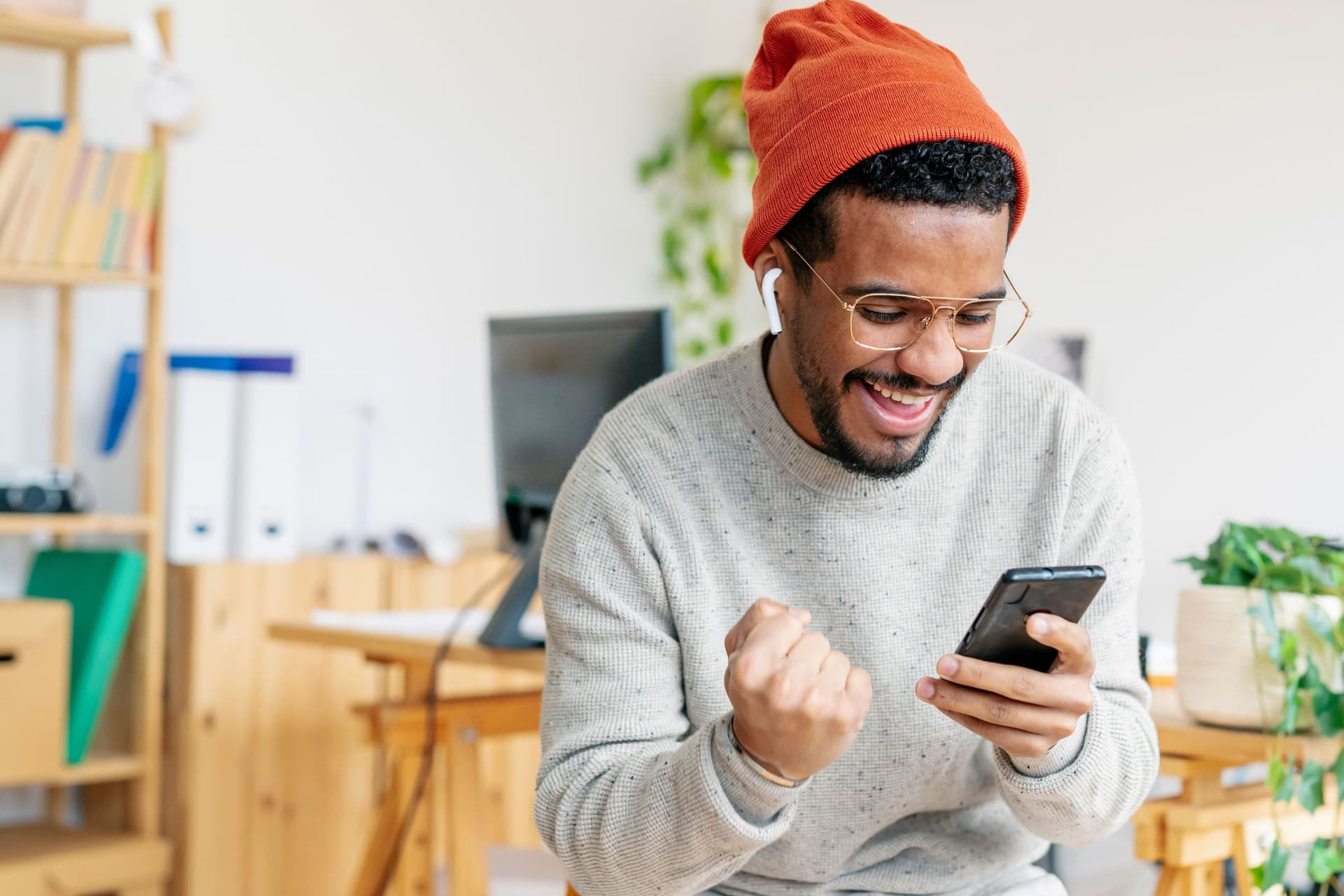 Junger Mann mit Mütze und Ohrhörern schaut lachend auf ein Smartphone und ballt die Faust