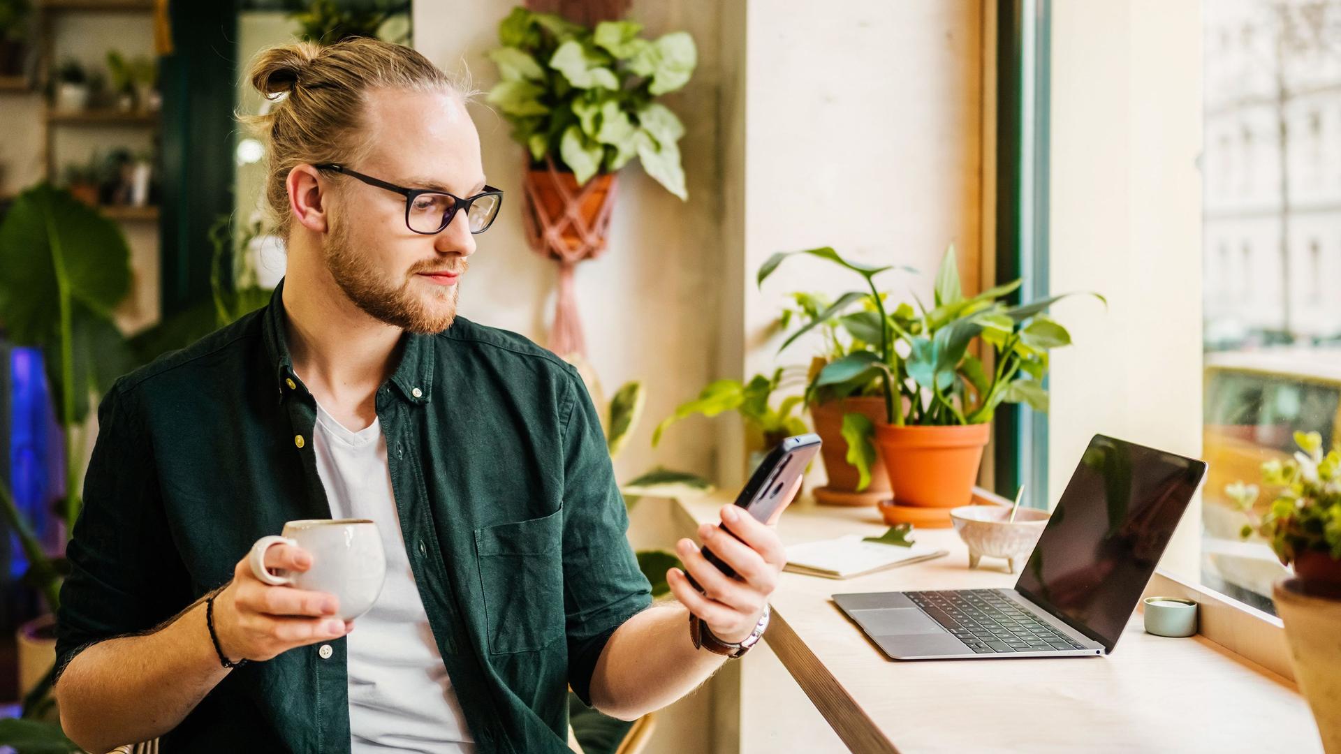 Junger Mann sitzt mit Laptop im Cafe und schaut auf sein Smartphone, während er in der rechten Hand eine Tasse hält.