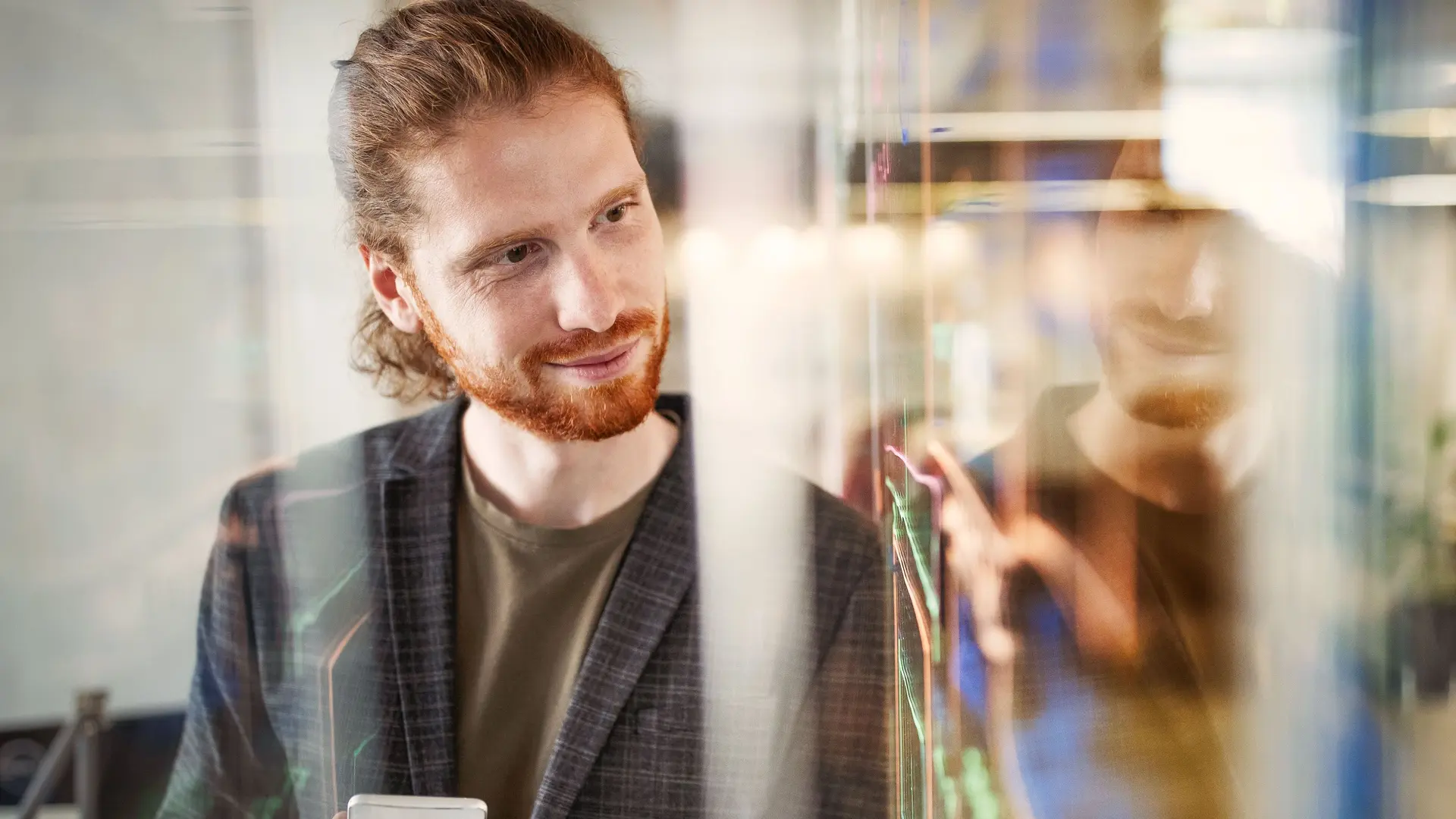 Junger Mann lehnt sich mit Stift in der Hand an eine beschriebene Glasscheibe im Büro an.