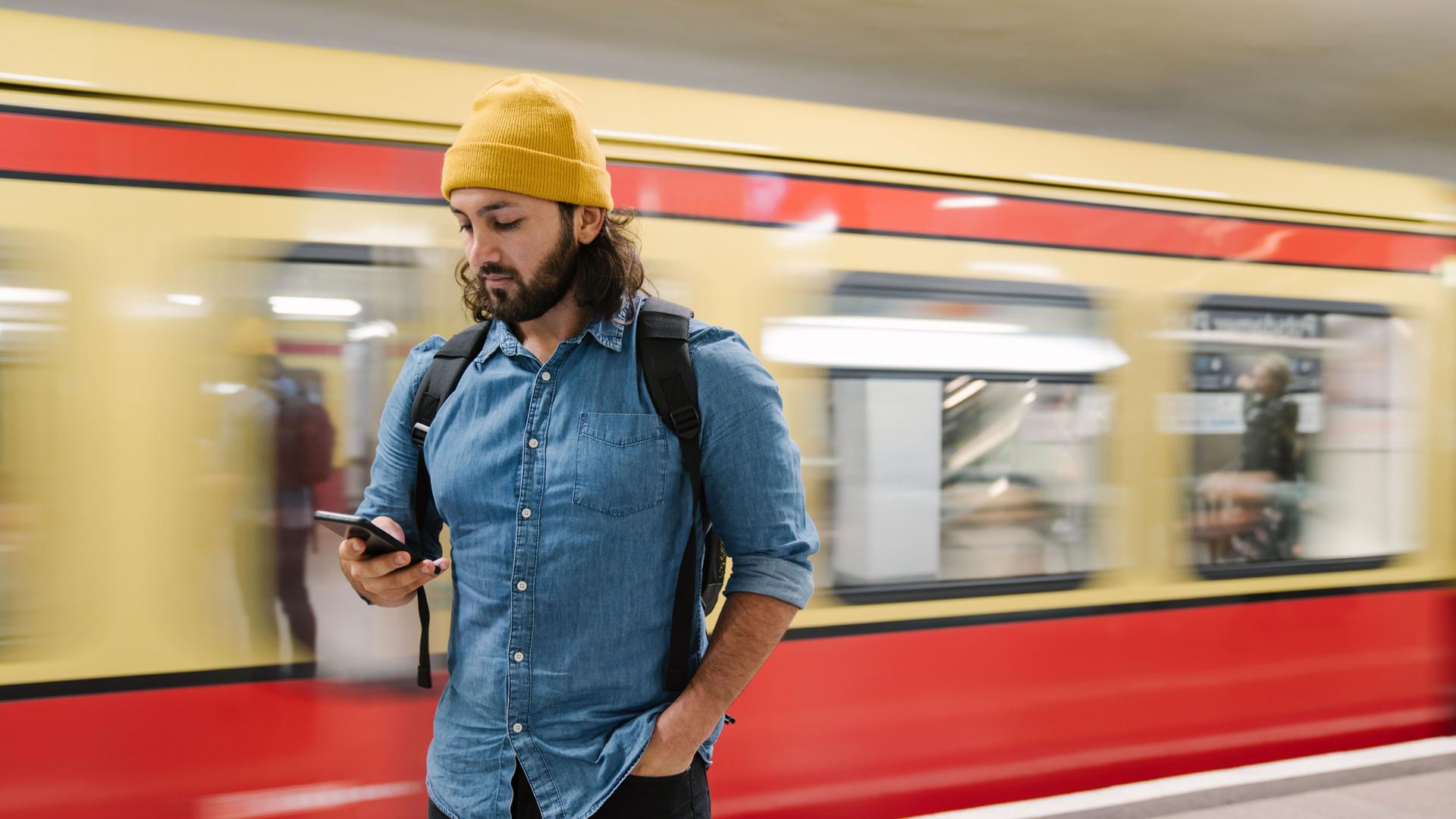 Ein Mann steht auf einem S/U-Bahnhof und blickt auf sein Mobiltelefon. Hinter ihm fährt eine U-Bahn vorbei.