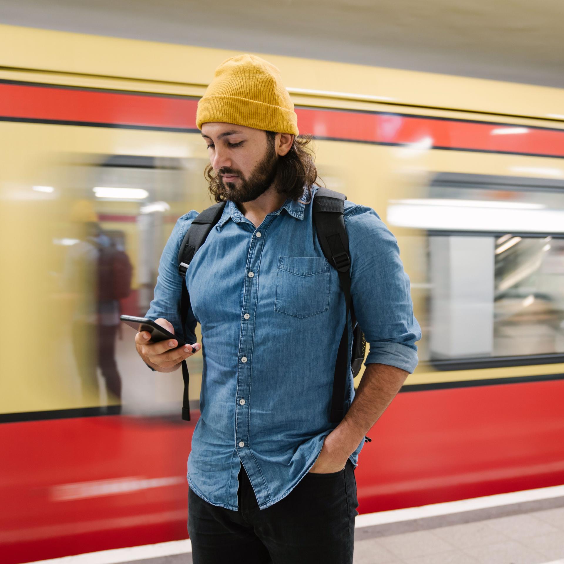 Ein Mann steht auf einem S/U-Bahnhof und blickt auf sein Mobiltelefon. Hinter ihm fährt eine U-Bahn vorbei.