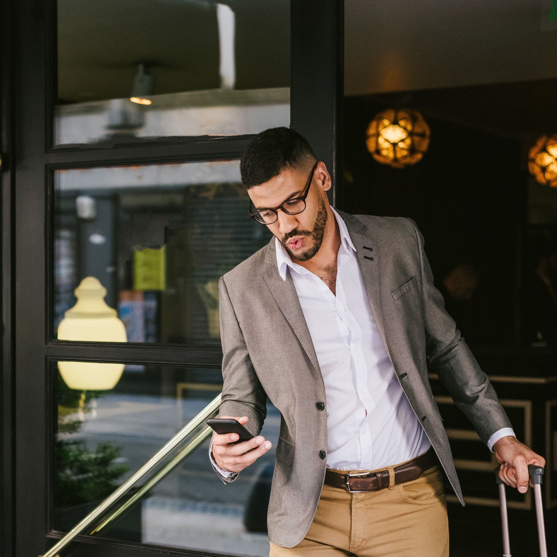 Junger Mann mit kurzen, dunklen Haaren, Brille und Bart zieht mit der einen Hand einen Rollkoffer und hält in der anderen ein Smartphone, während er ein Gebäude durch eine von mehreren verglasten Türen verlässt.
