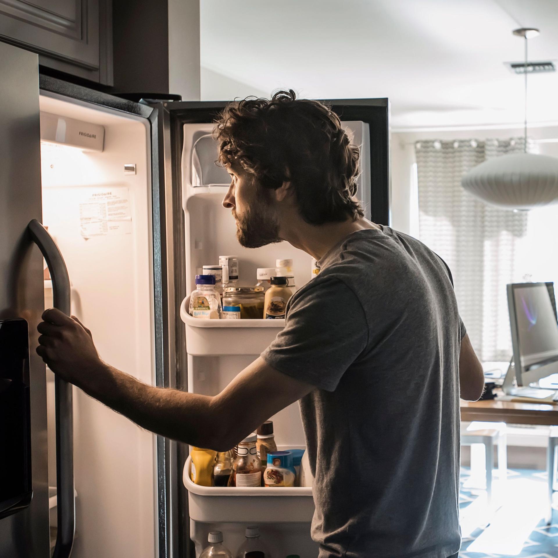 Ein Mann steht vor einem geöffneten Kühlschrank und schaut hinein.