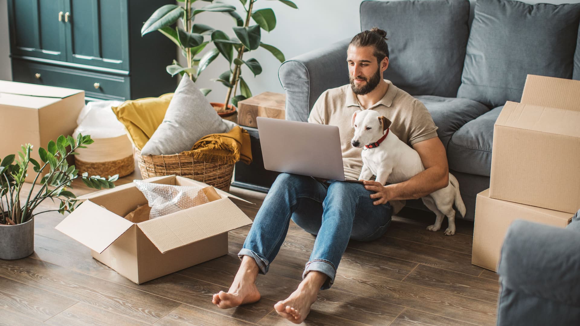 Junger Mann sitzt beim Umzug vor einem Sofa und hat einen Laptop und seinen Hund auf dem Schoß.