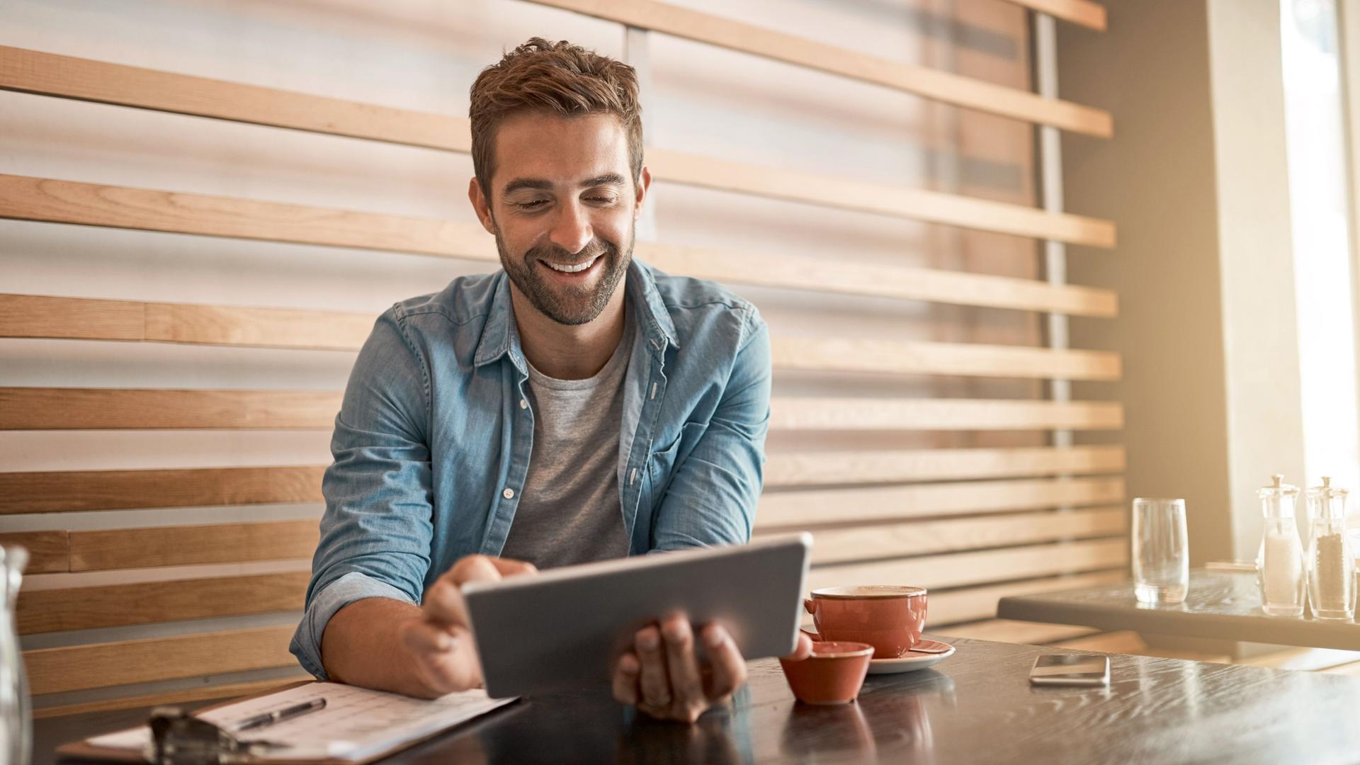 Junger Mann sitzt lachend in einem Cafe und arbeitet an seinem Tablet
