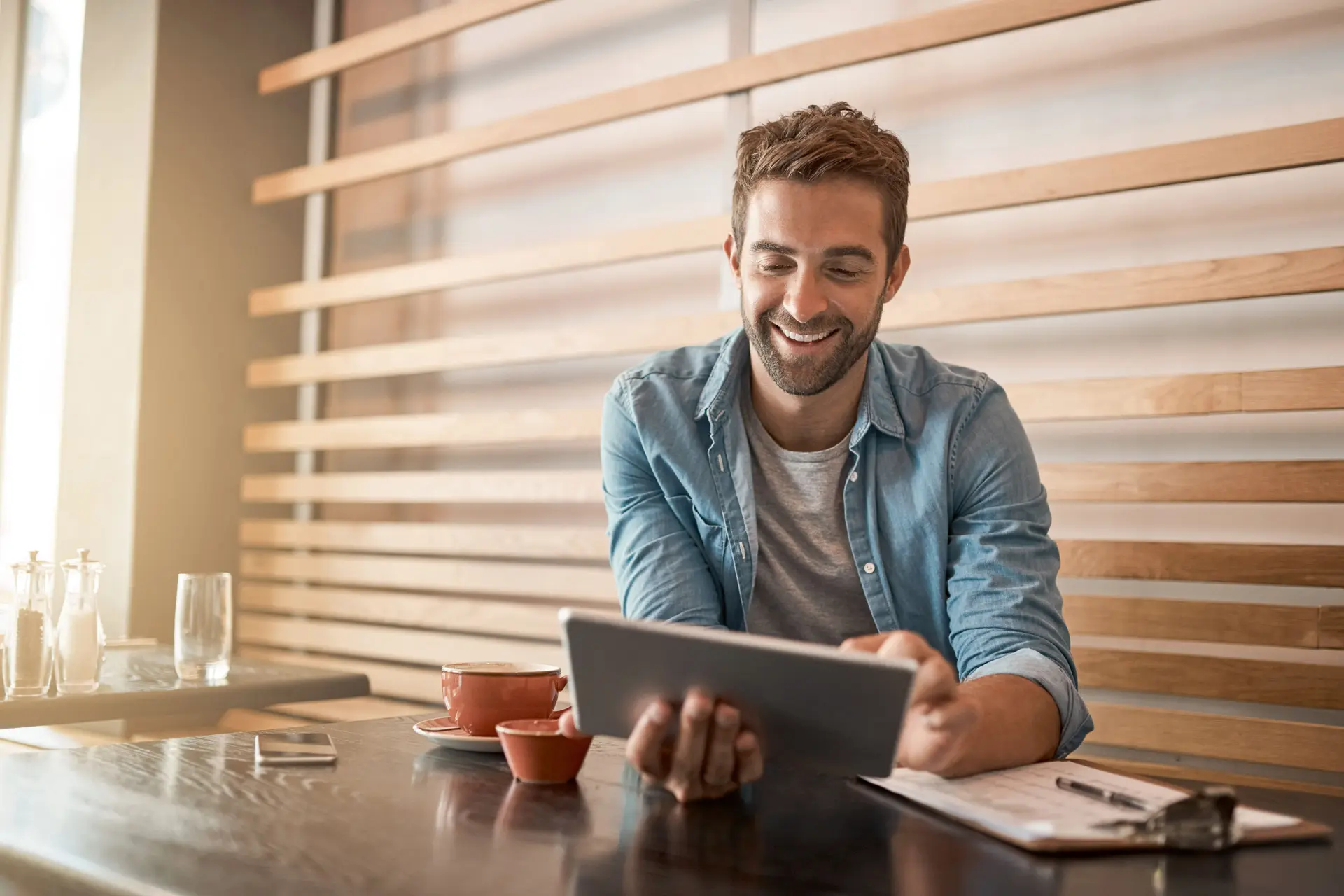 Junger Mann sitzt lachend in einem Cafe und arbeitet an seinem Tablet