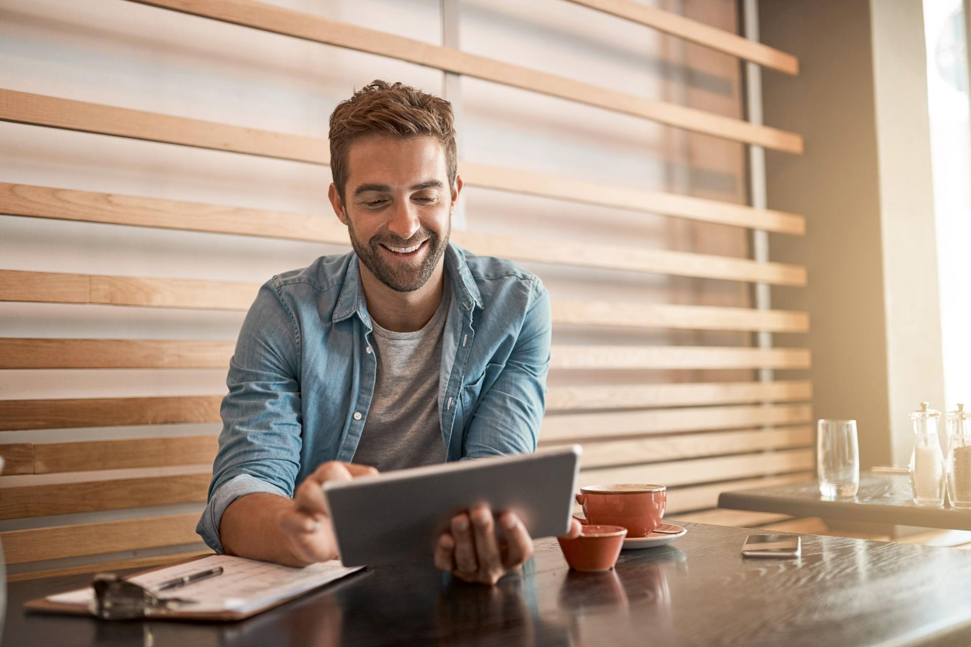Junger Mann sitzt lachend in einem Cafe und arbeitet an seinem Tablet