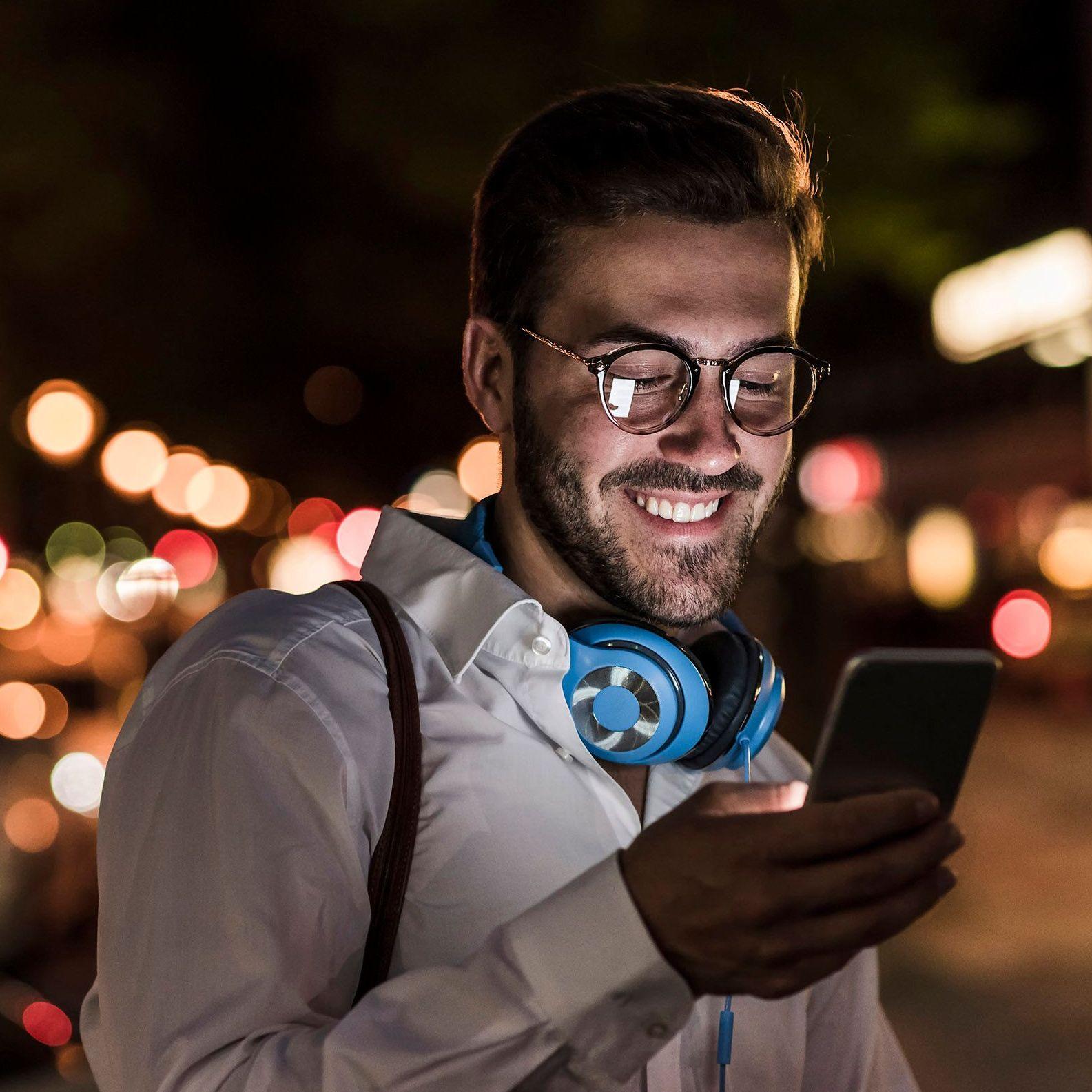 Ein junger Mann im weißen Hemd und türkisen Kopfhörern um den Hals, schaut glücklich auf den Display seines Smartphones. Es ist Abend und sein Gesicht wird vom Displaylicht angeleuchtet.