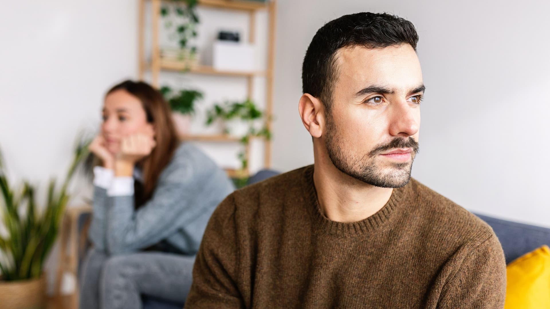 Junges Paar sitzt distanziert auf einem Sofa. Einer junger Mann sitzt vorne und blickt zur Seite, während die Frau im Hintergrund in die andere Richtung blickt.