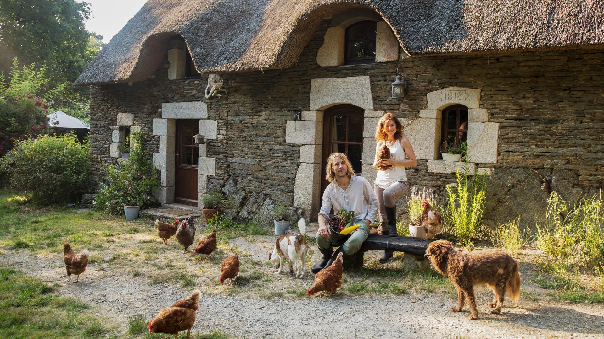 Junges Paar vor einem alten Bauernhof mit Reetdach in der Bretagne. Um sie herum sind Hunde und Hühner.