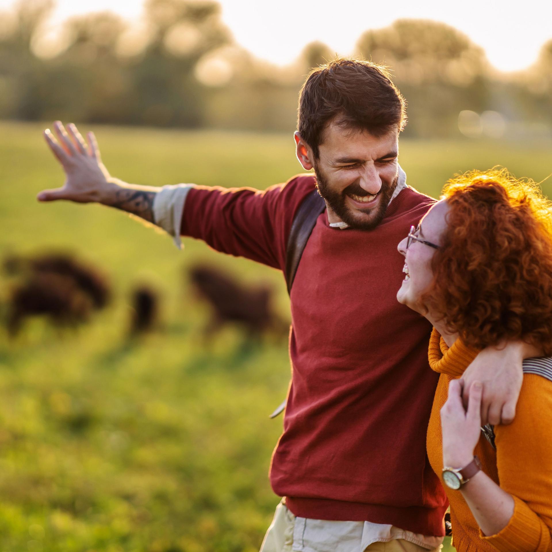 Junges Paar läuft Arm in Arm in der Natur und guckt sich dabei lächelnd an. Im Hintergrund scheint die Sonne auf grüne Wiesen und Ziegen.