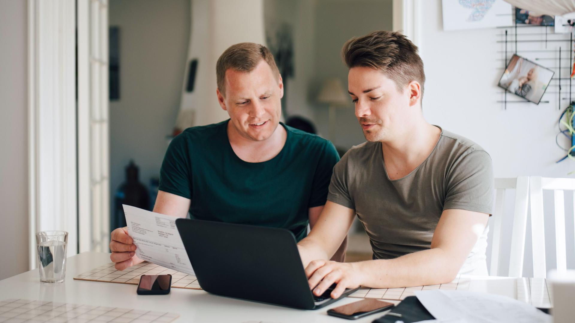 Zwei Männer an einem Küchentisch mit Laptop zahlen Rechnungen online.