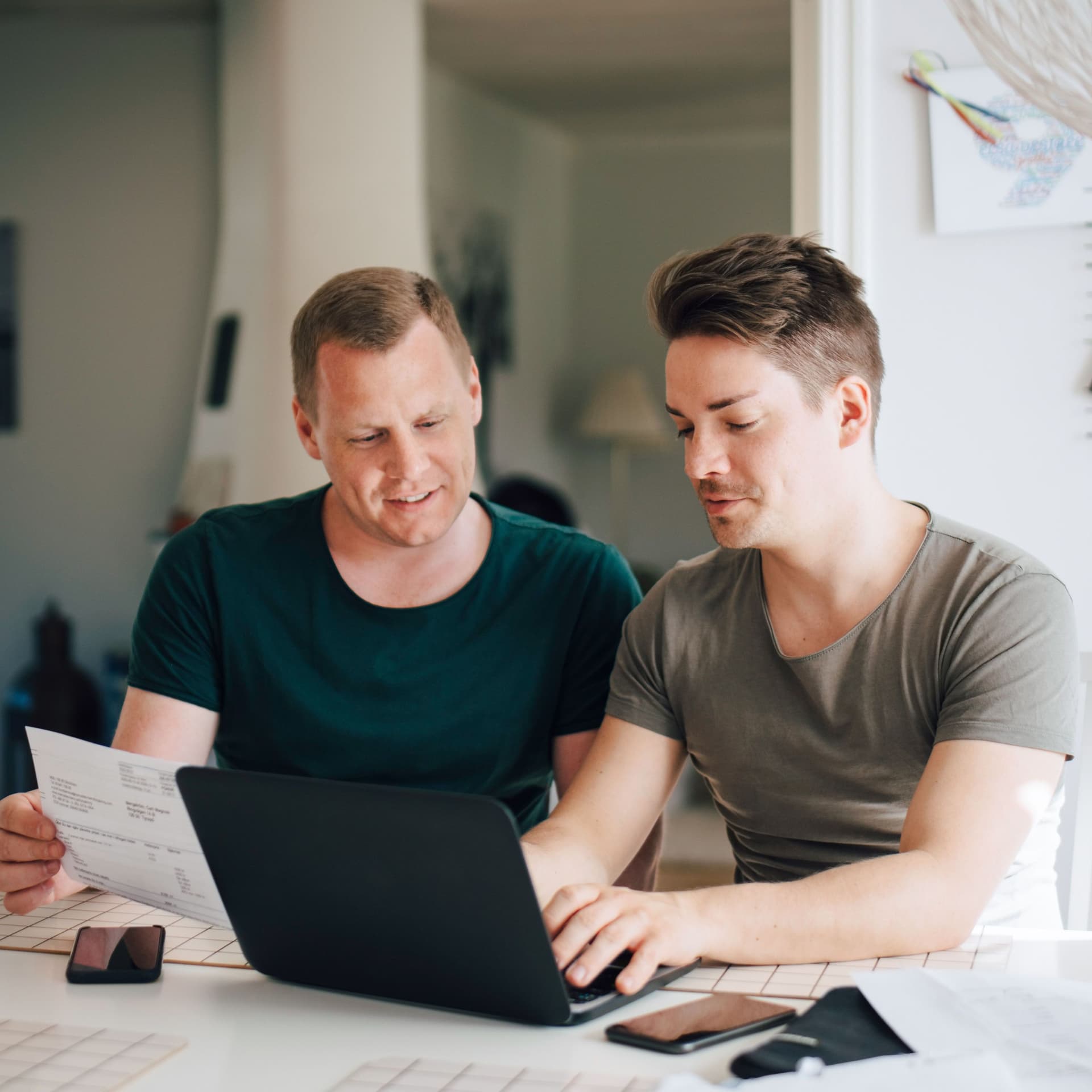 Zwei Männer an einem Küchentisch mit Laptop zahlen Rechnungen online.