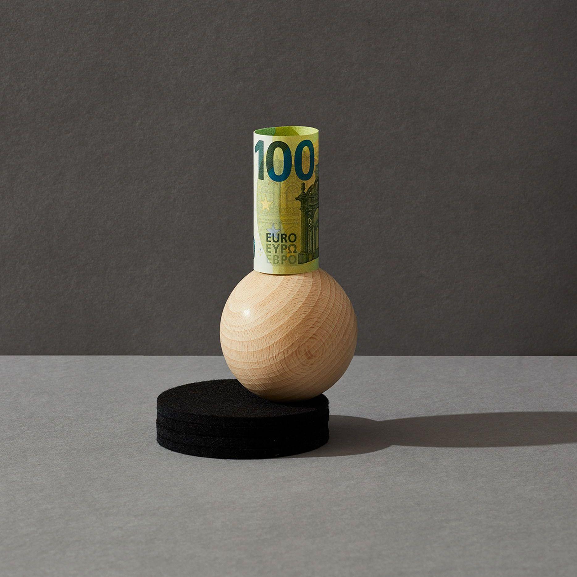 Eine gerollte 100-Euro-Banknote steht senkrecht aufgerichtet auf einer Holzkugel vor grauem Hintergrund. 