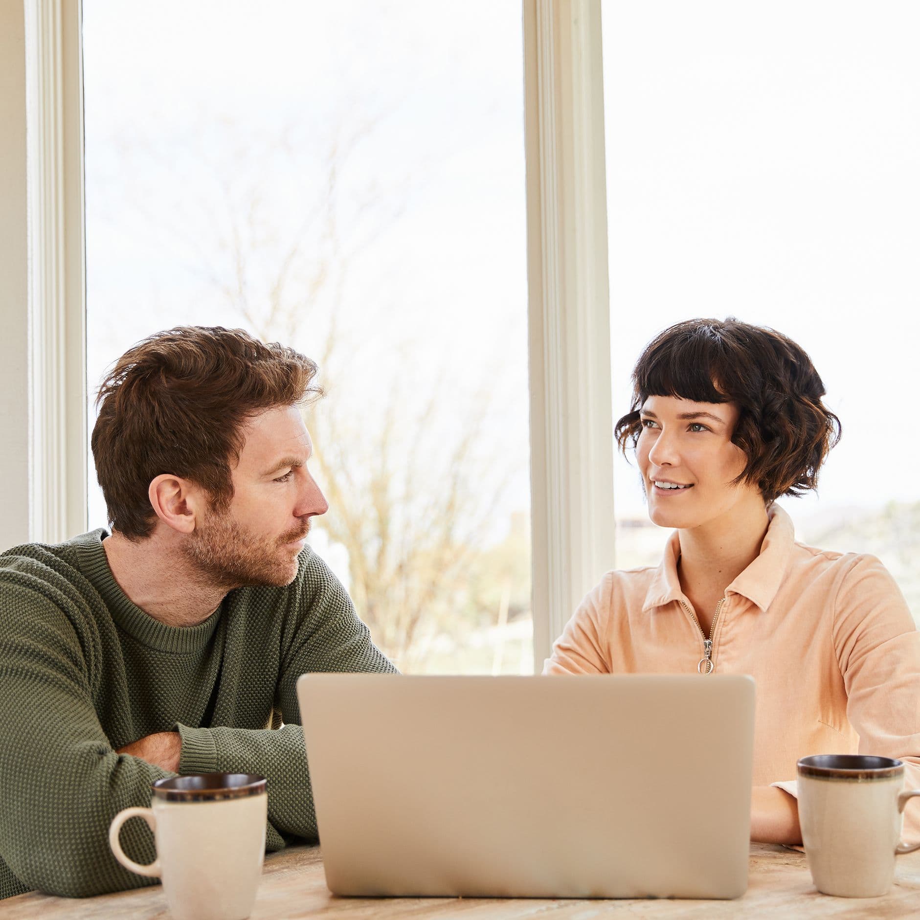 Ein Mann und eine Frau sitzen an einem Tisch vor einem Laptop und unterhalten sich. Beide haben Kaffeetassen vor sich, und durch das große Fenster im Hintergrund fällt Tageslicht in den Raum.