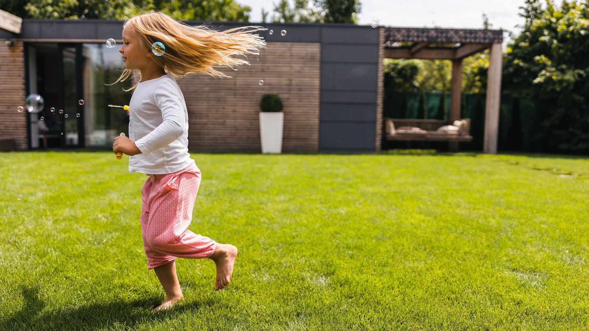 Blondes Mädchen rennt barfuß über Rasen in einem Garten. Um sie herum sind Seifenblasen.