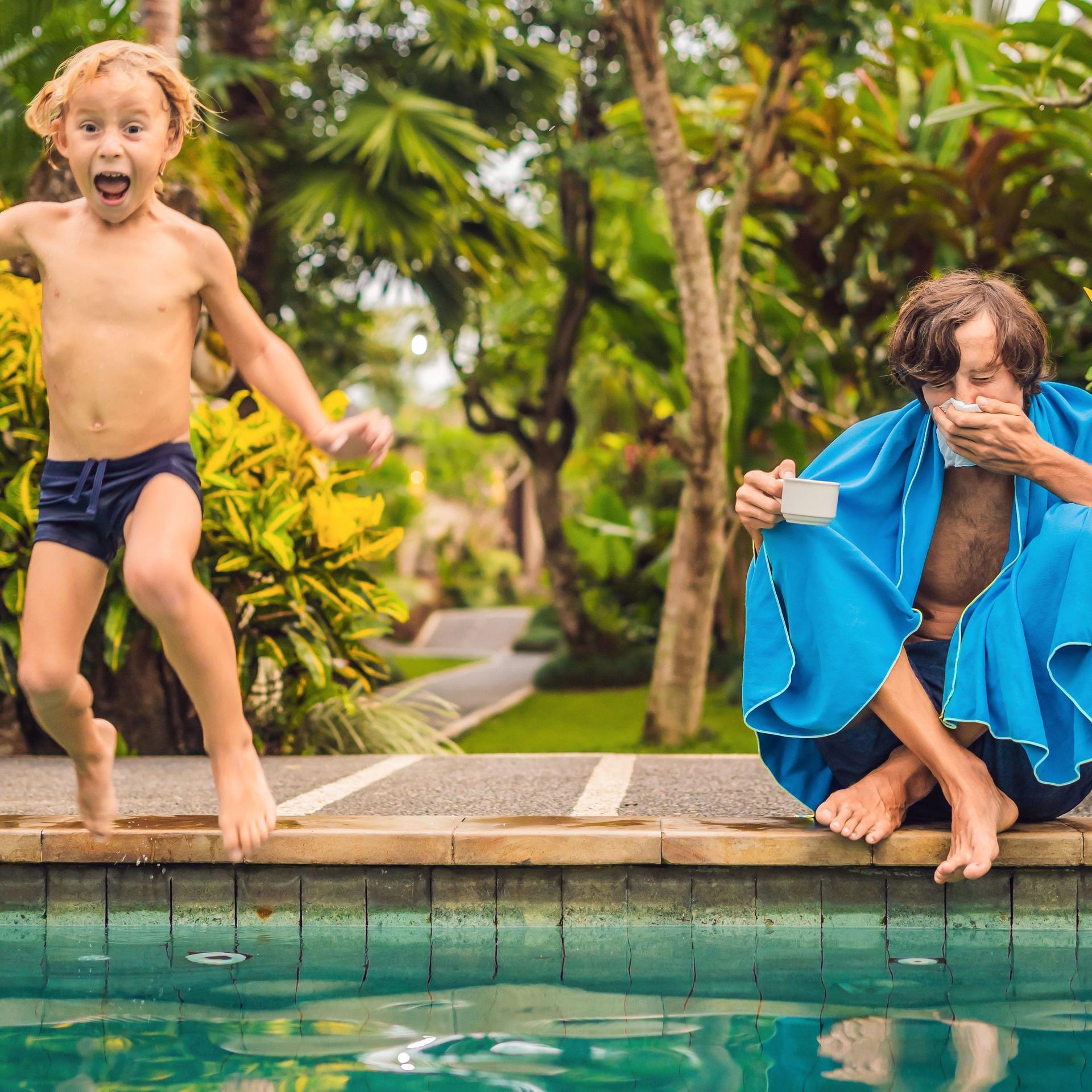 Verschnupfter Vater mit Kind am Pool. Das Kind springt lachend in den Pool während sich der Vater die Nase putzt.
