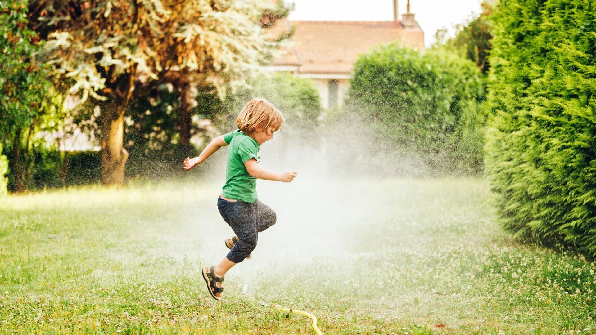 Kind springt über einen Gartensprinkler auf einer Wiese.