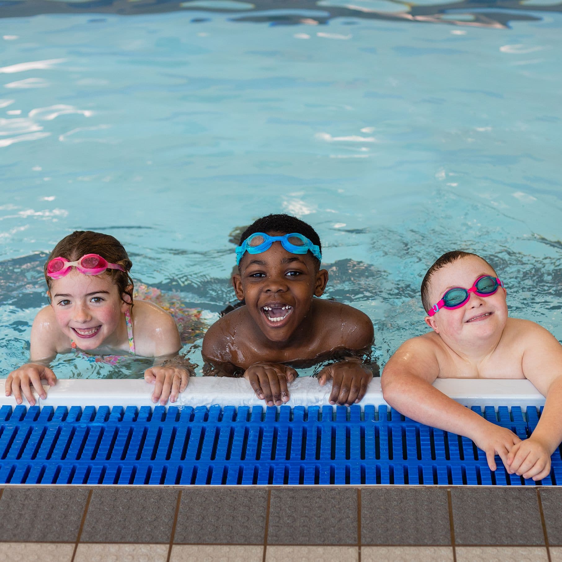 Drei fröhliche Kinder mit bunten Schwimmbrillen halten sich am Beckenrand eines Hallenbads fest und lächeln in die Kamera.