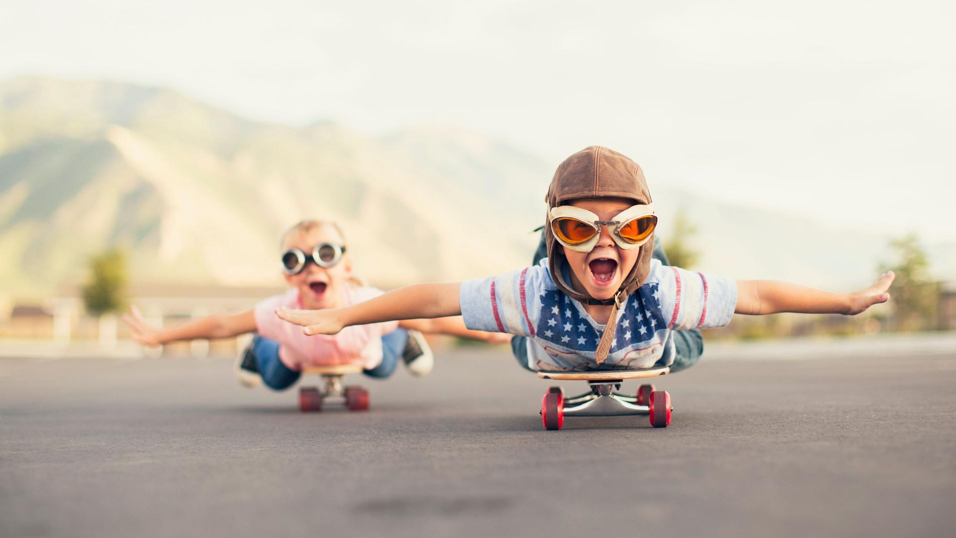 Zwei Kinder fahren bäuchlings auf einem Skateboard und strecken dabei die Arme aus als ob sie fliegen würden.