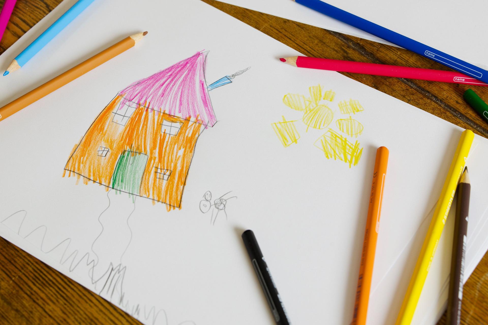 Ein Kinderzeichnung liegt mit Buntstiften auf einem Holztisch. Die Zeichnung zeigt ein Haus und eine Sonne.