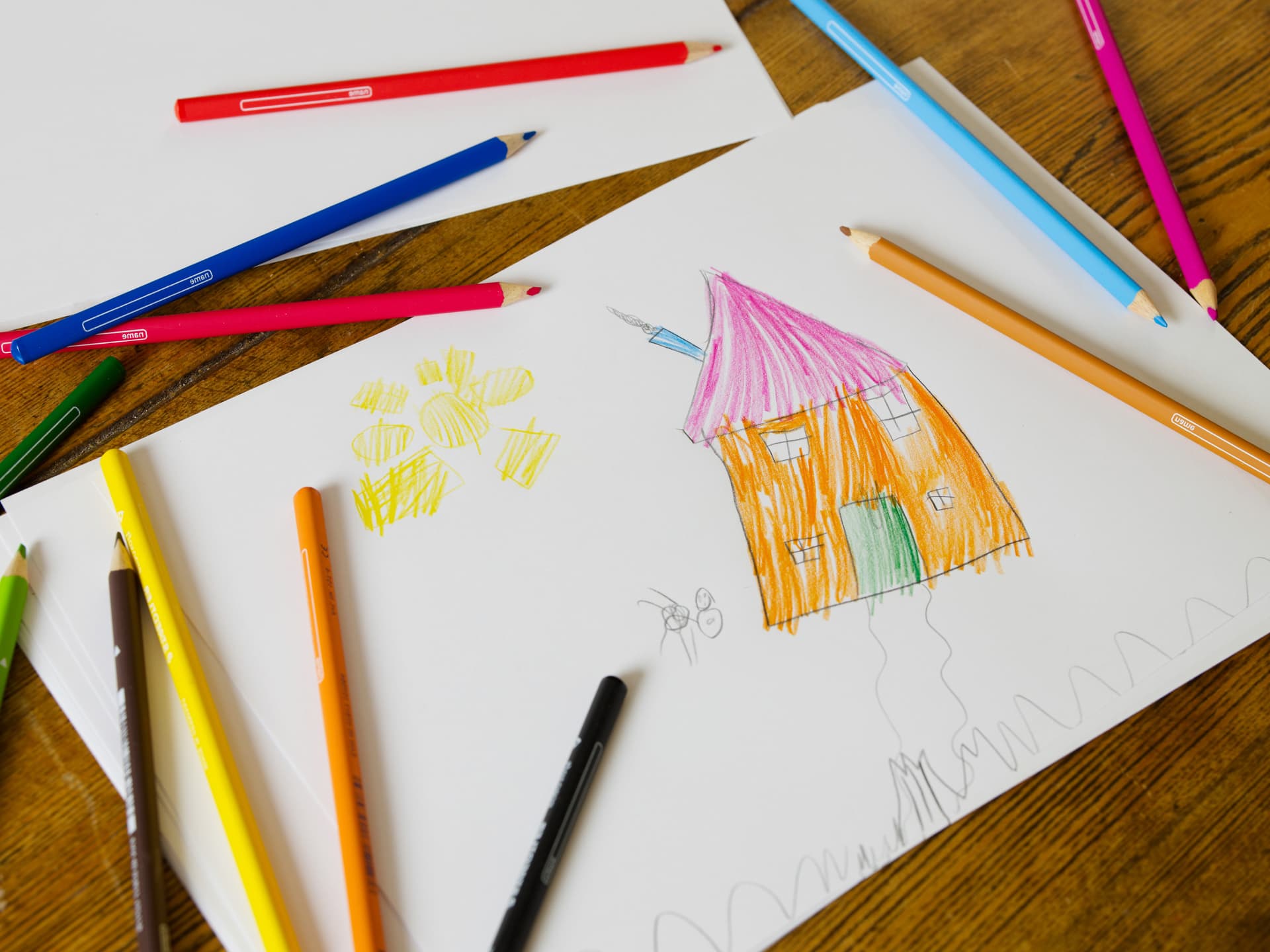 Ein Kinderzeichnung liegt mit Buntstiften auf einem Holztisch. Die Zeichnung zeigt ein Haus und eine Sonne.
