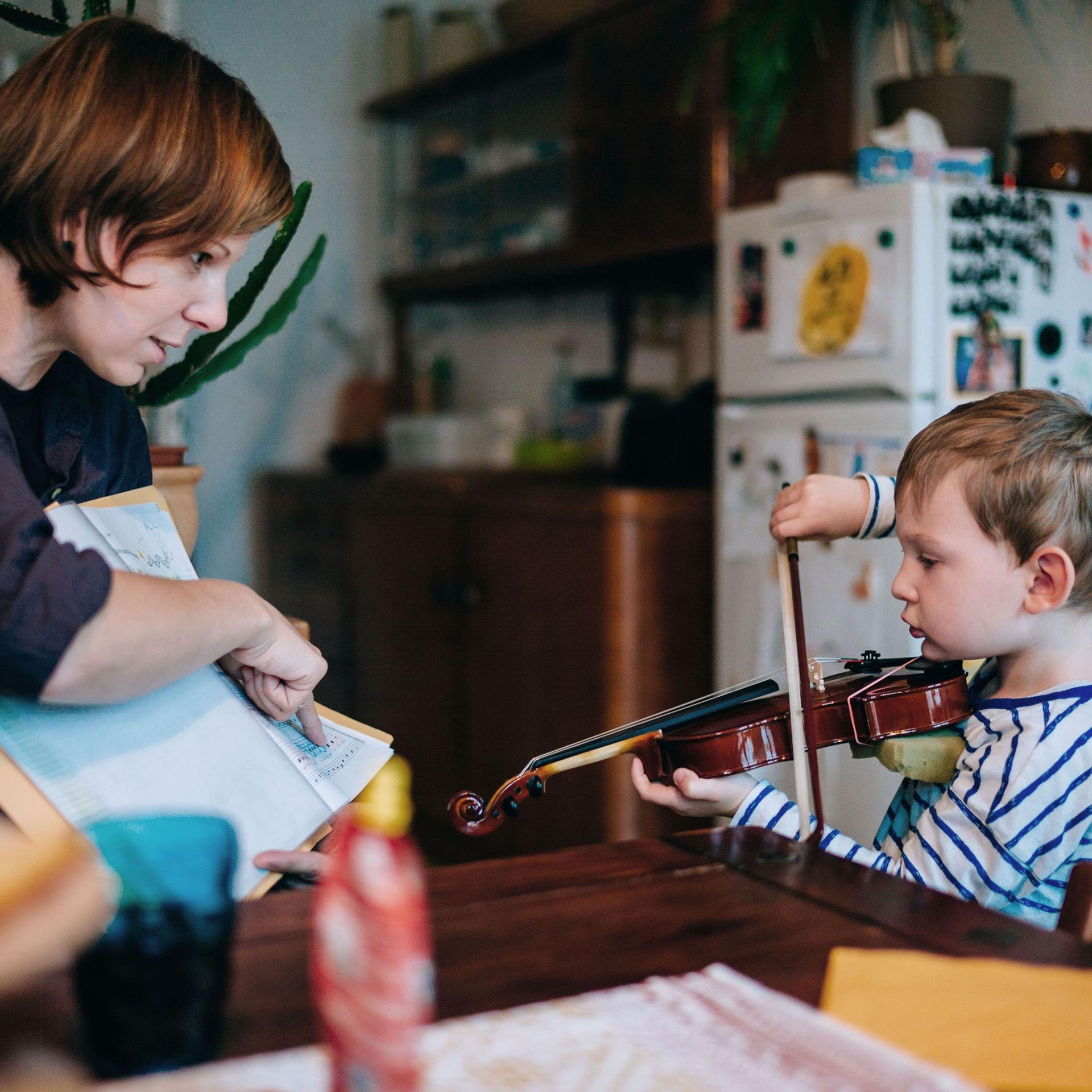 Ein Kind wird zuhause von einer Frau an der Geige unterrichtet. Sie zeigt dem Kind die Noten, während es spielt. Beide schauen konzentriert. 