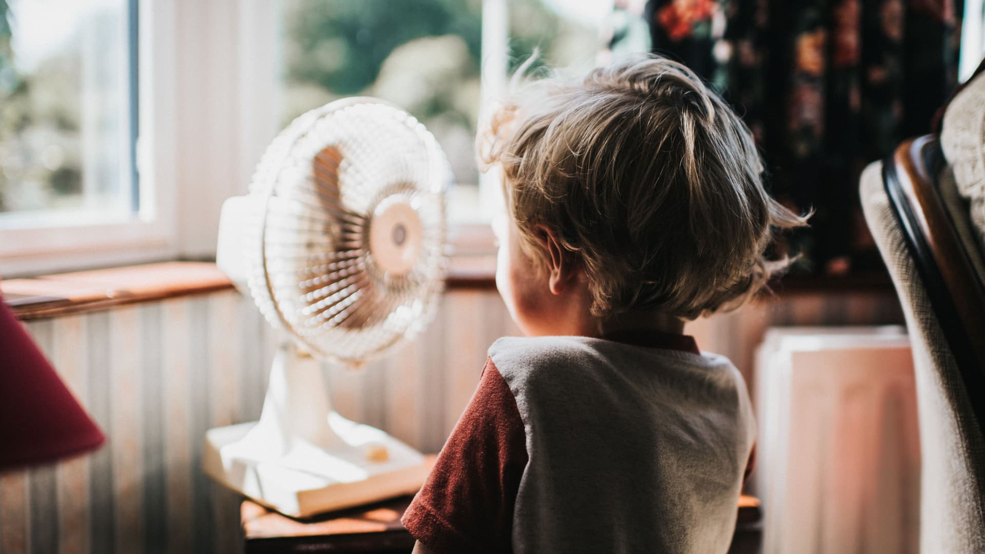 Ein Kleinkind steht vor einem kleinen Ventilator. Die Haare wehen dabei im Wind. Im Hintergrund sind Fenster zu sehen.