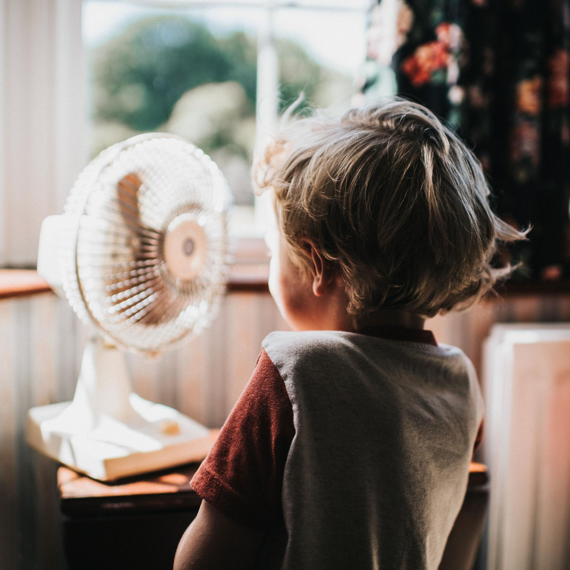 Ein Kleinkind steht vor einem kleinen Ventilator. Die Haare wehen dabei im Wind. Im Hintergrund sind Fenster zu sehen.