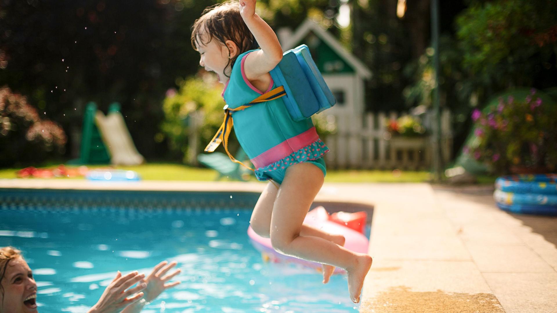 Ein Mädchen im Kindergartenalter springt mit Schwimmweste in einen Pool. Ein Mann steht im Wasser und streckt ihr die Arme entgegen.