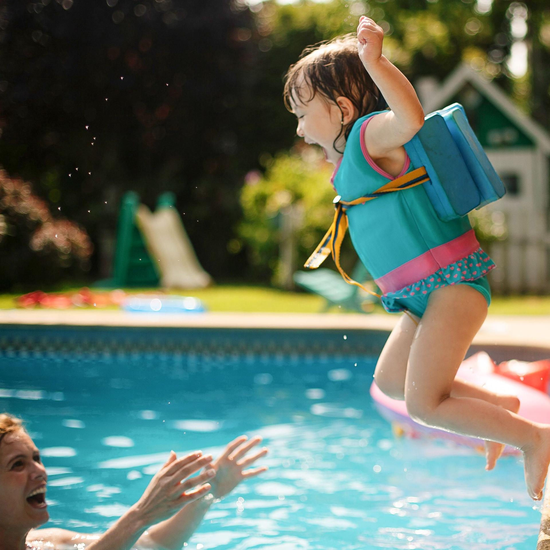 Ein Mädchen im Kindergartenalter springt mit Schwimmweste in einen Pool. Ein Mann steht im Wasser und streckt ihr die Arme entgegen.