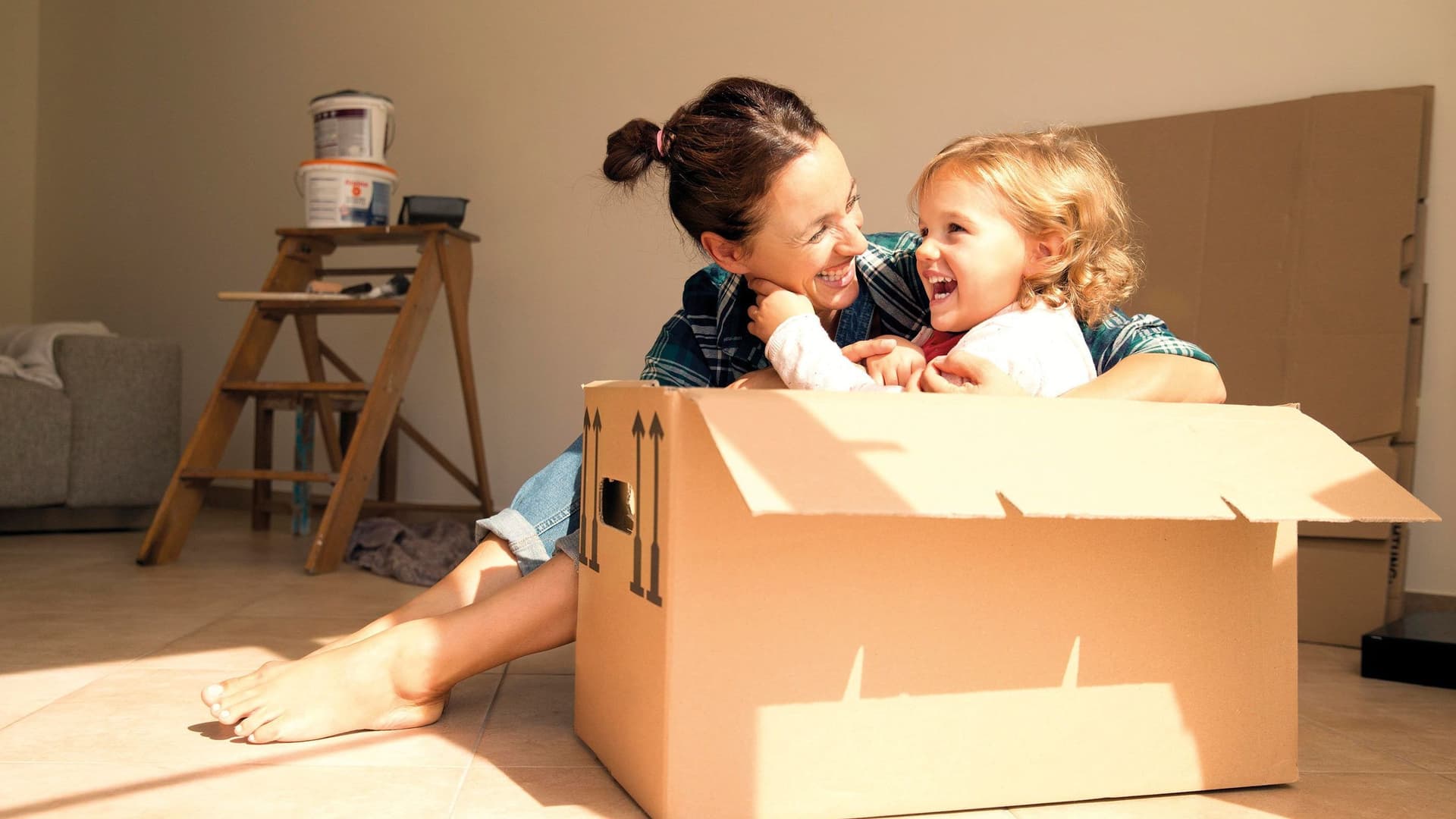 Ein kleines Kind im Kindergartenalter sitzt in einem Karton in einer Umzugswohnung. Neben ihm sitzt die Mutter und umarmt es.