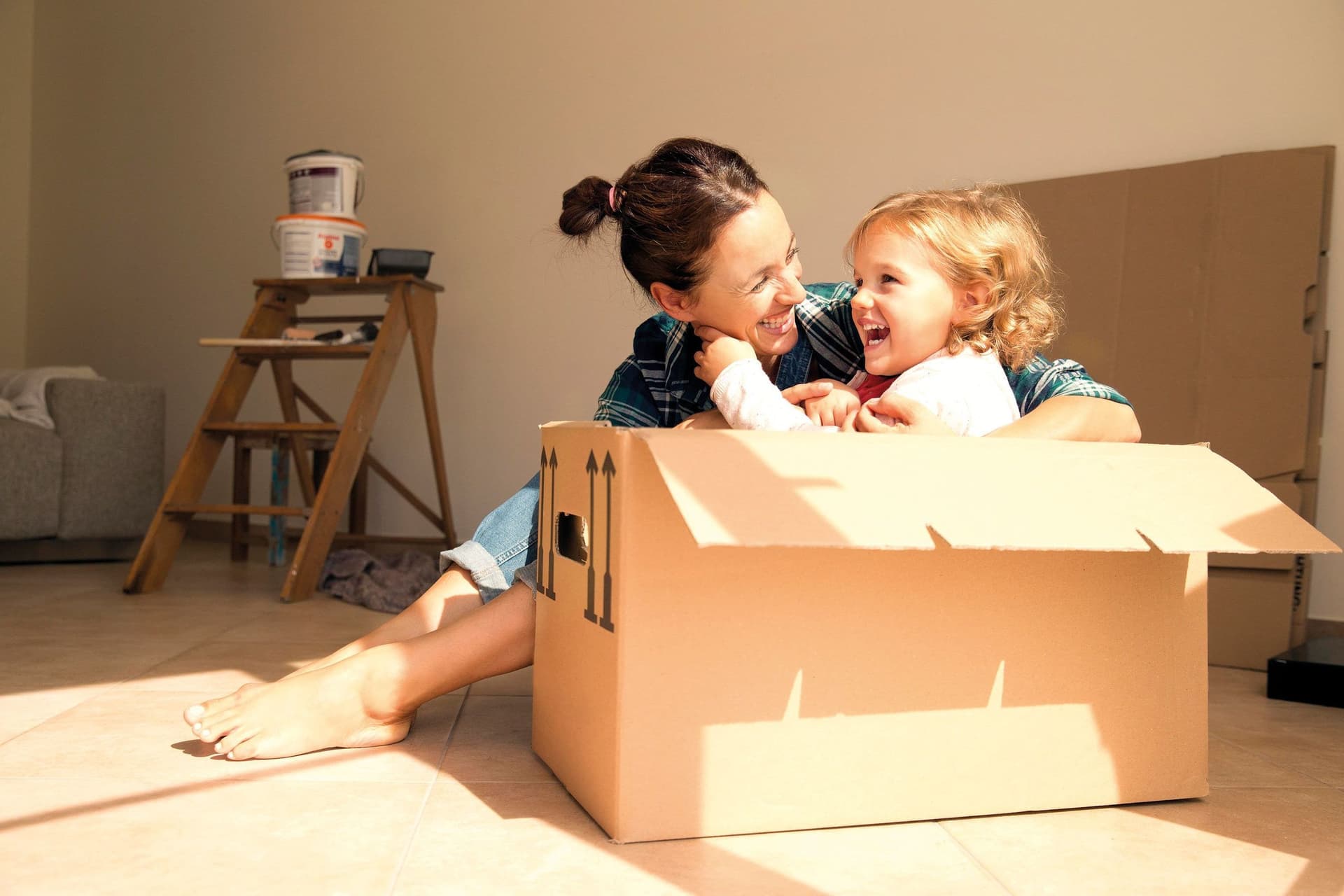Ein kleines Kind im Kindergartenalter sitzt in einem Karton in einer Umzugswohnung. Neben ihm sitzt die Mutter und umarmt es.