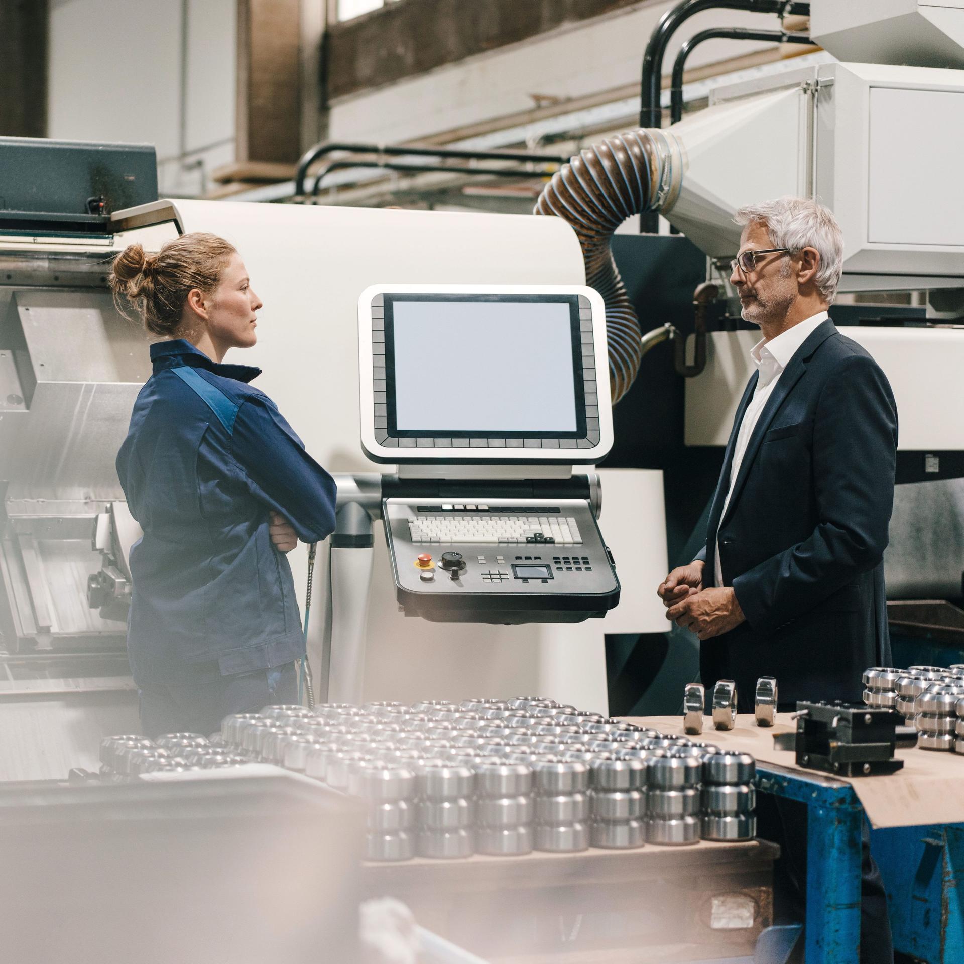 Frau und Mann stehen vor einer Maschine mit Bildschirm in einer Produktionshalle.