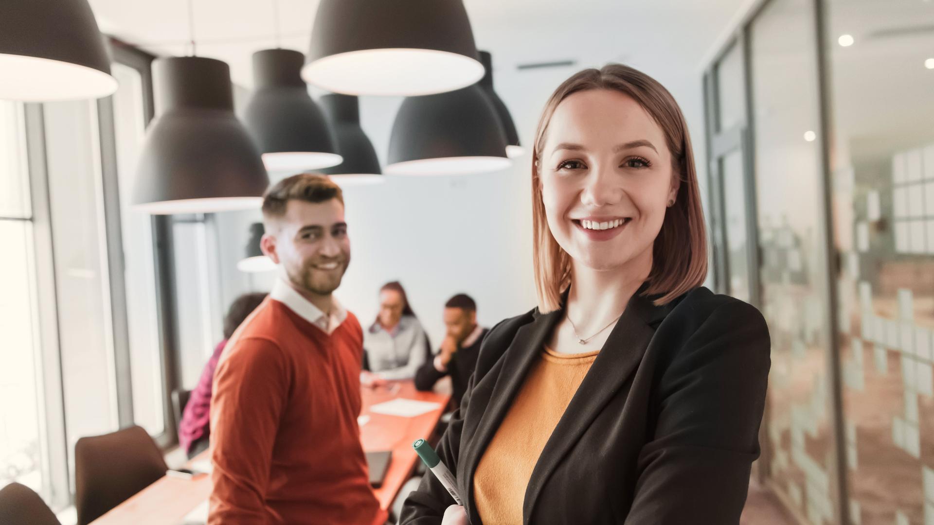 Porträt einer glücklichen Geschäftsfrau und eines Geschäftsmannes in einem modernen Büro. Sie lächeln und blicken in die Kamera. Ein Team arbeitet im Hintergrund. 