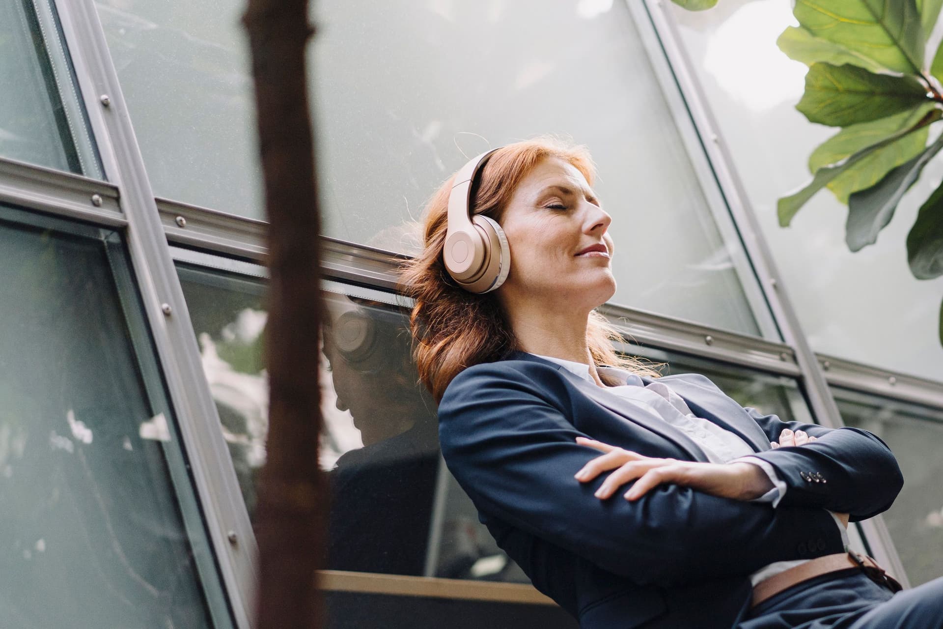 Frau mit Kopfhörern sitzt lächelnd und mit geschlossenen Augen auf einer Bank vor einem modernen Bürogebäude. Sie hat die Arme verschränkt und lehnt sich nach hinten.