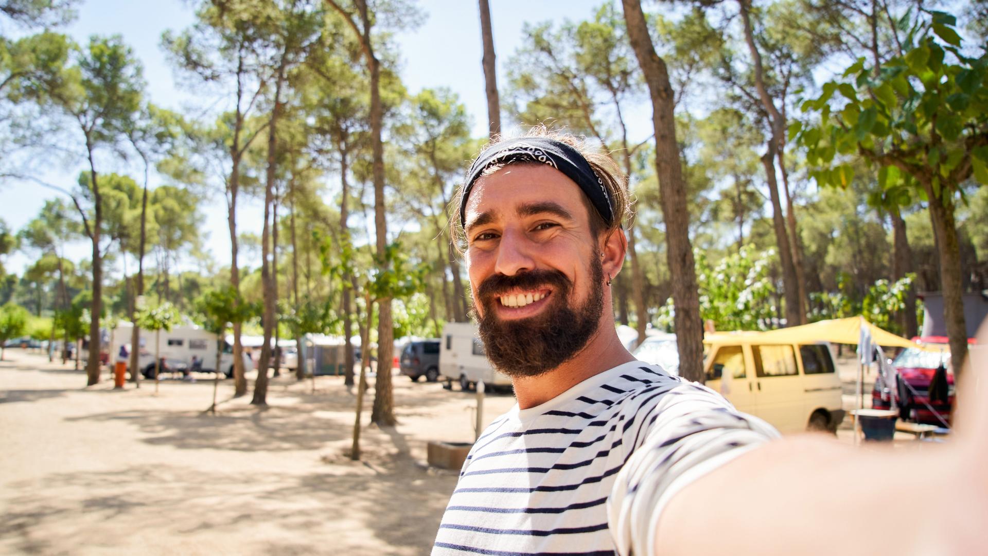 Ein fröhlicher junger Mann mit Vollbart und gestreiftem T-Shirt macht ein Selfie auf einem Campingplatz mit Wohnmobilen in einem Kiefernwald.