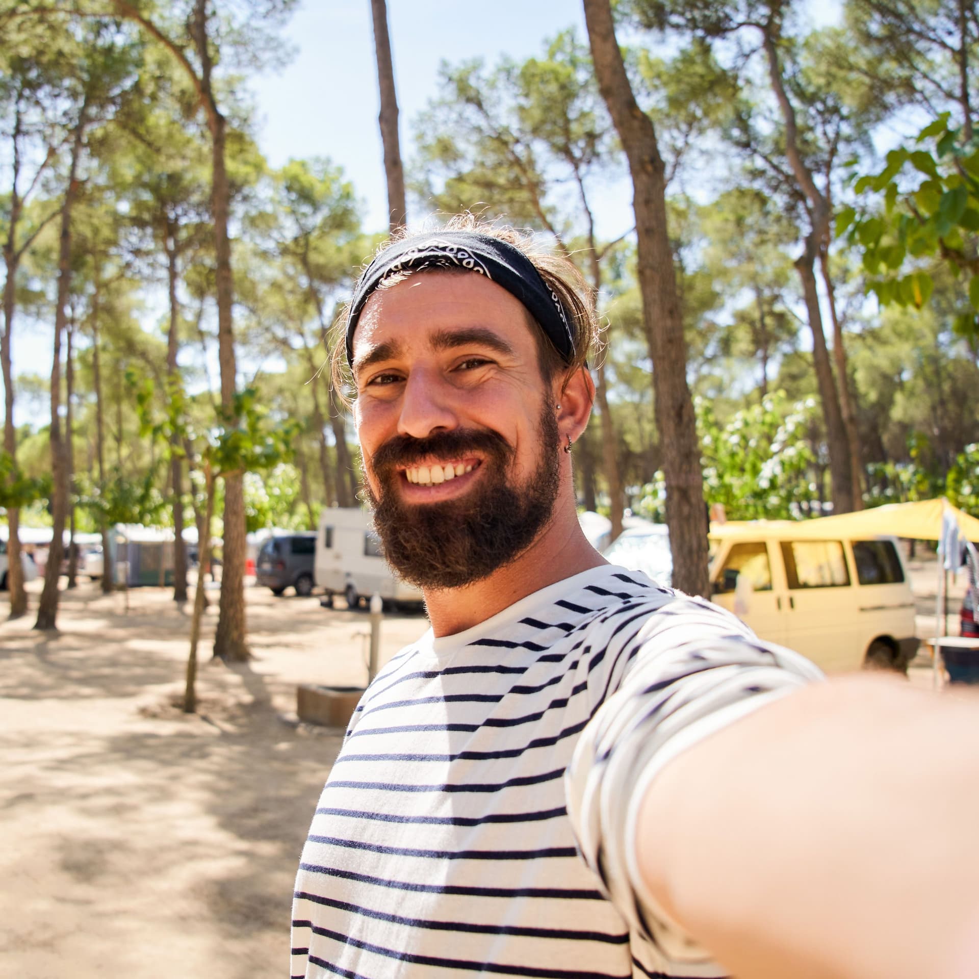 Ein fröhlicher junger Mann mit Vollbart und gestreiftem T-Shirt macht ein Selfie auf einem Campingplatz mit Wohnmobilen in einem Kiefernwald.